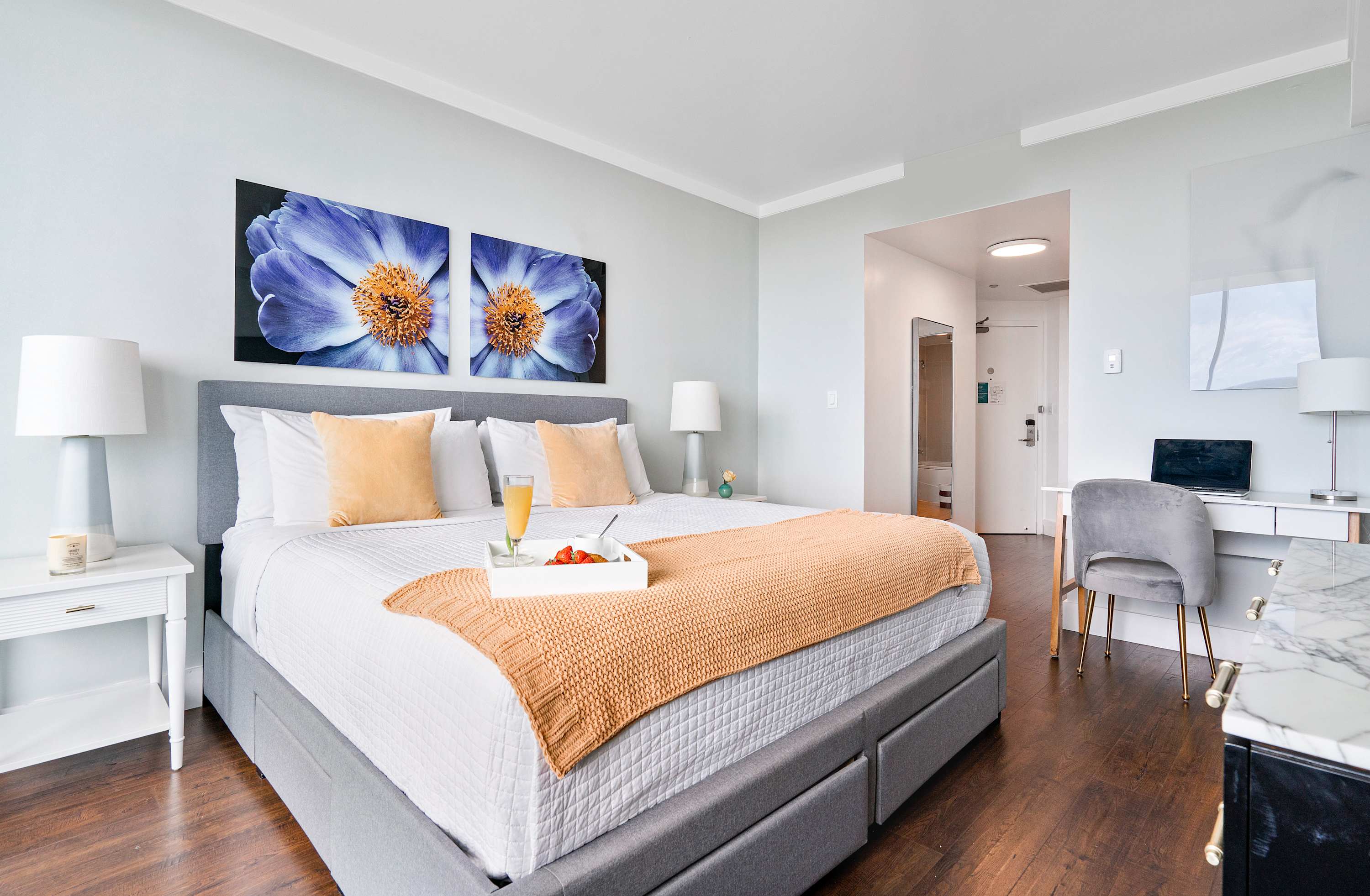 A spacious bedroom with a large bed, two blue floral artworks, and a dedicated workspace with a white desk and gray chair, offering a glimpse into the hallway and bathroom entrance.