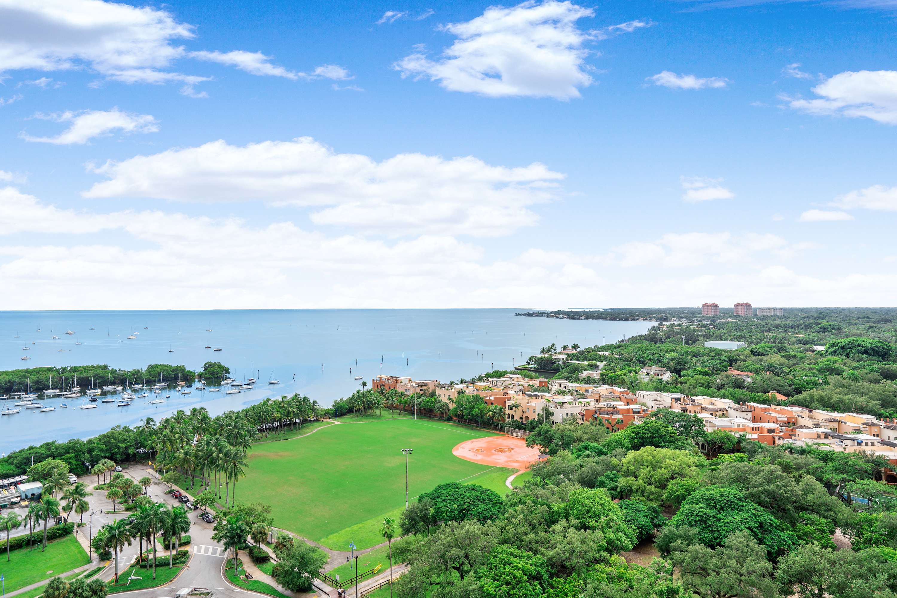 A high-angle view captures a wide expanse of calm blue bay dotted with boats, adjacent to a large green park with a baseball field, and residential buildings under a bright, cloudy sky.
