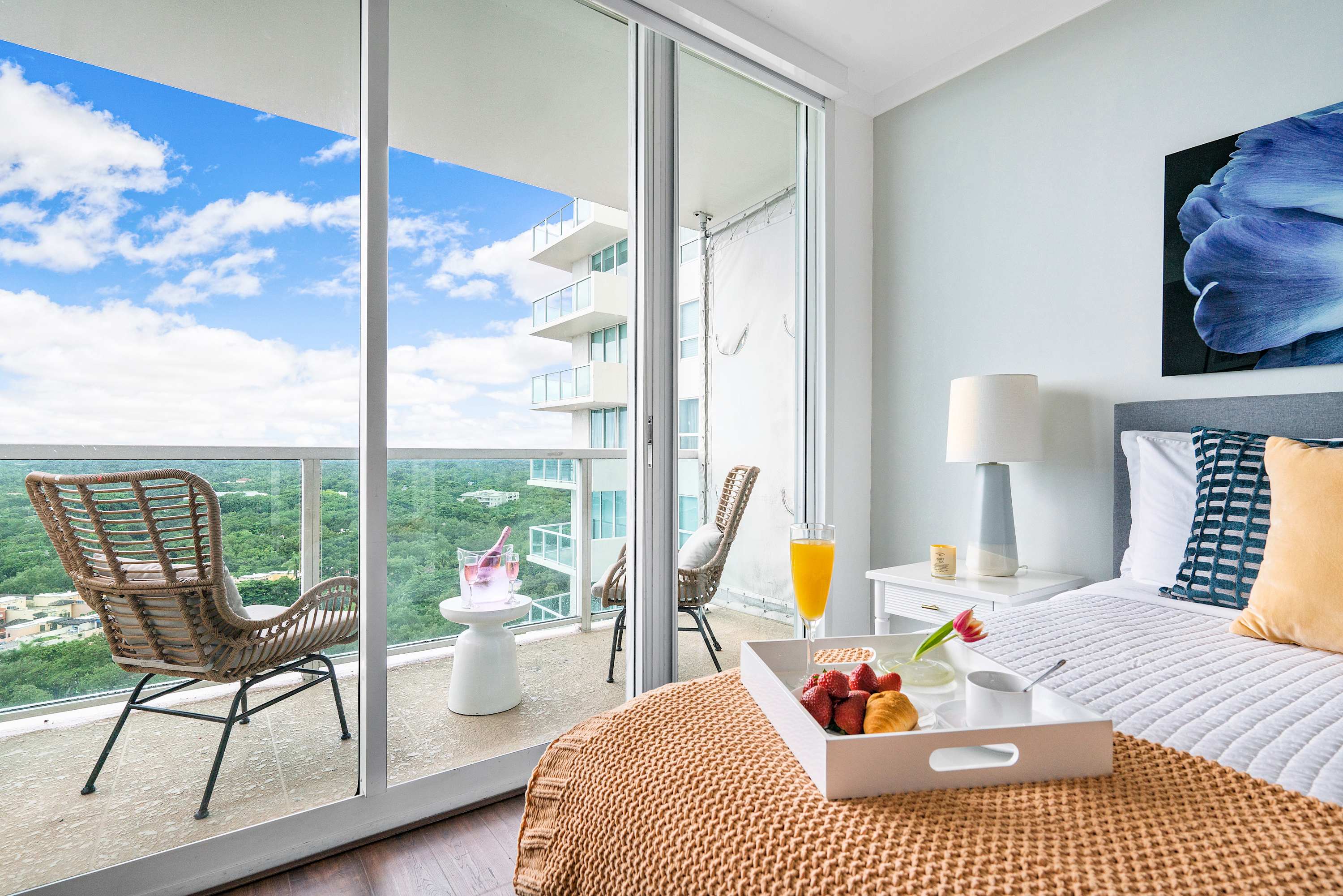 A cozy bedroom with a white bed, gold throw, and a breakfast tray, featuring a large sliding glass door that opens to a balcony with two wicker chairs and a view of lush greenery and distant buildings.