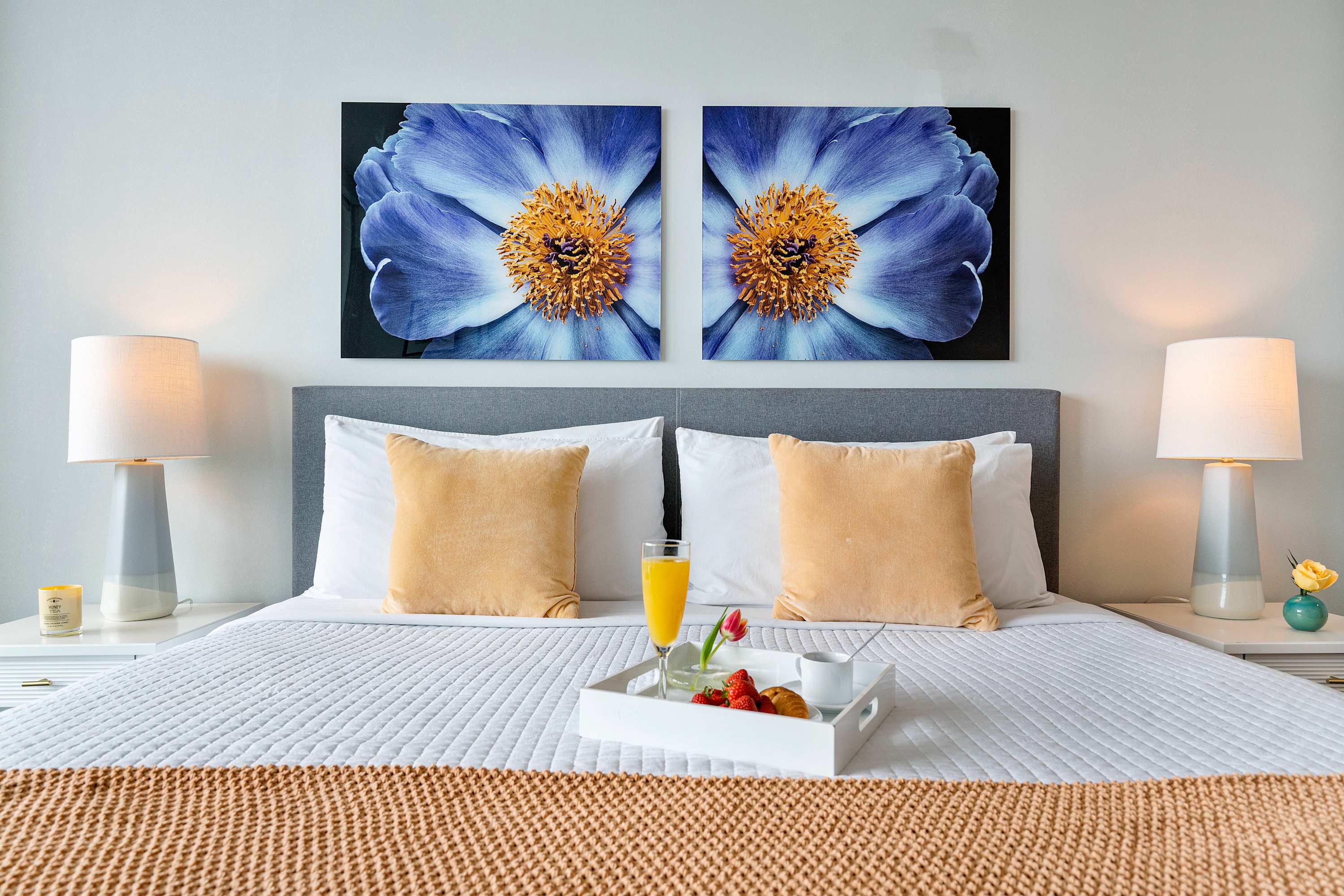 A neatly made bed with white linens and a gold throw is flanked by two white nightstands with lamps, featuring two large blue floral artworks above the gray headboard.