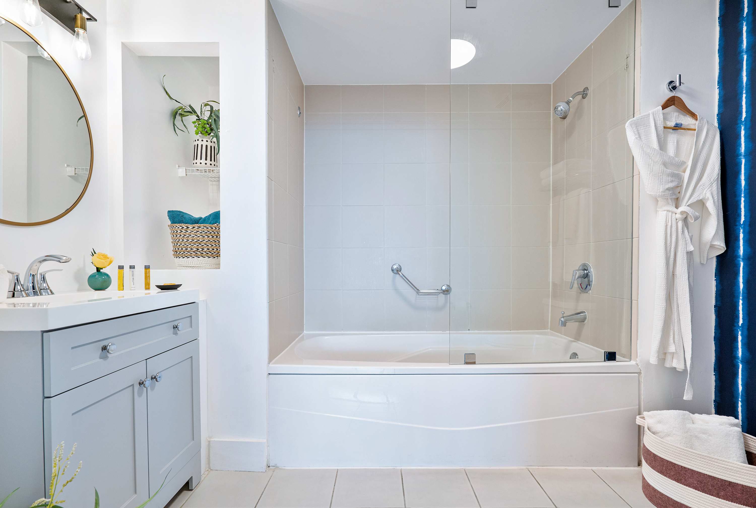 A clean and bright bathroom features a bathtub with a glass shower screen, a white vanity with a round mirror, and a white bathrobe hanging nearby, with blue patterned shower curtain visible.
