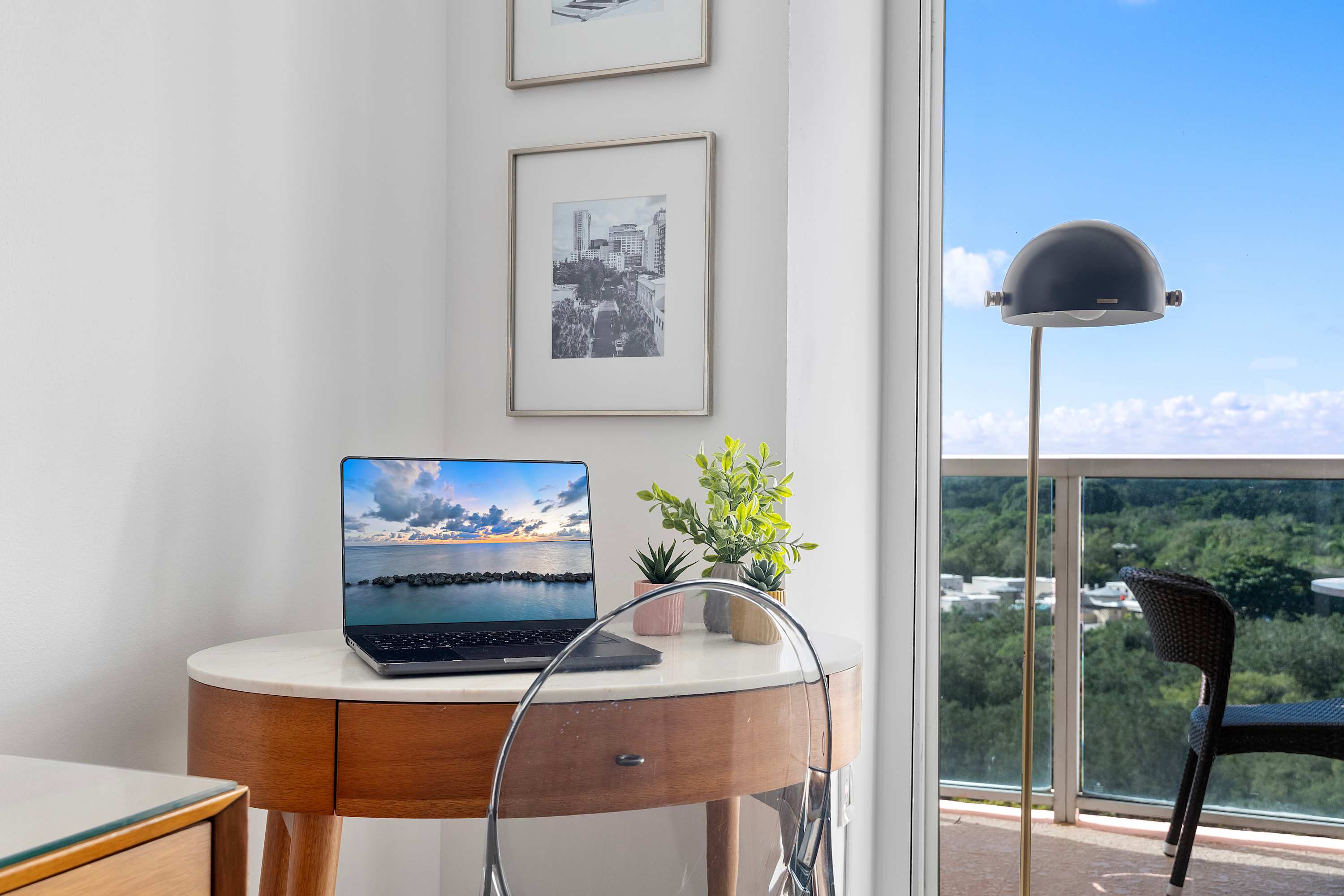Close-up of a small, round wooden desk with a laptop, a clear chair, and potted plants, positioned by a large window offering a bright outdoor view.