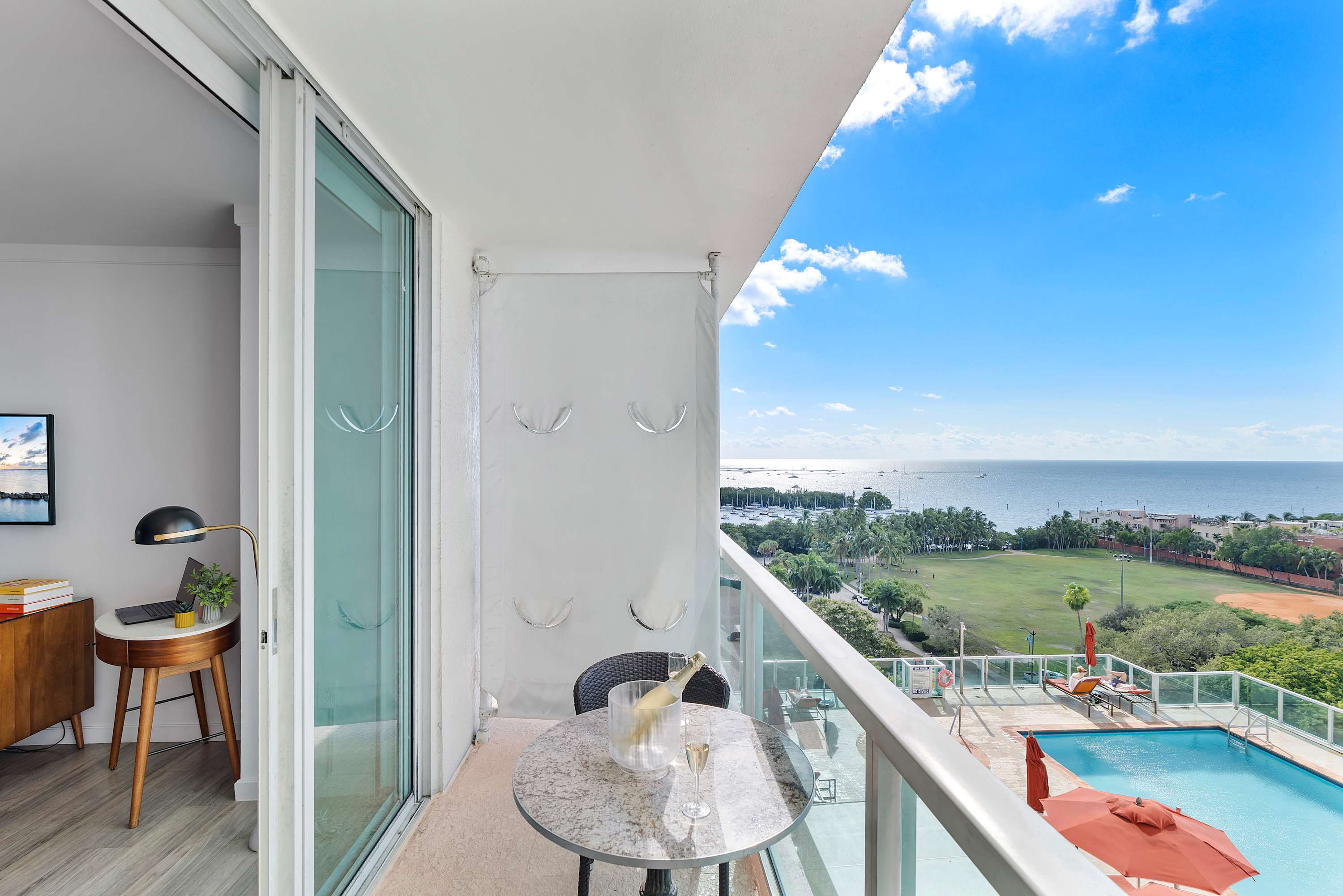 Balcony with a small table and two chairs, featuring a bottle of champagne in an ice bucket, overlooking a swimming pool and the expansive bay with boats.