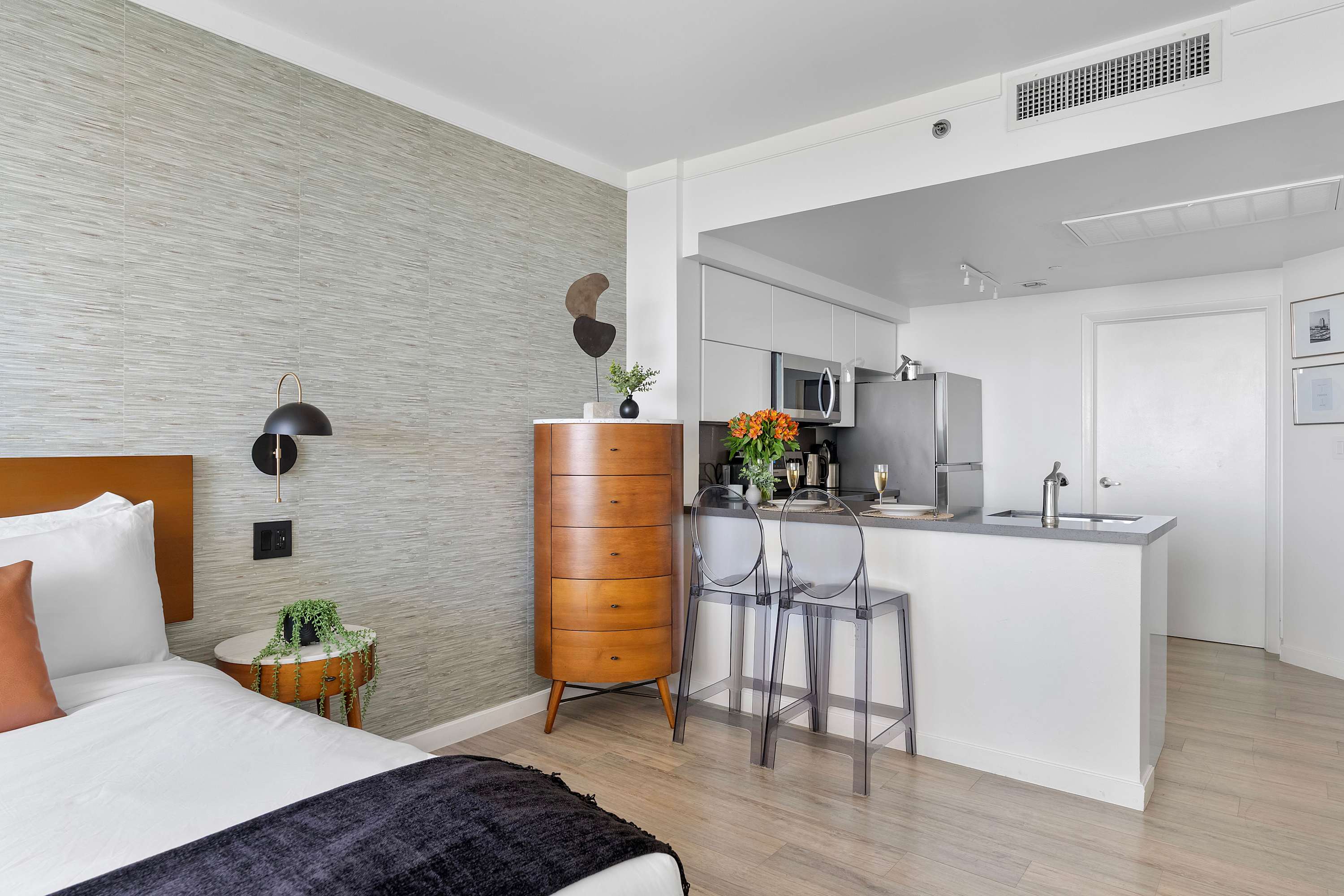 A bright studio apartment featuring a bed with a dark throw, a mid-century modern wooden dresser, and a view of the integrated kitchen bar area with clear bar stools. A textured accent wall adds visual interest.