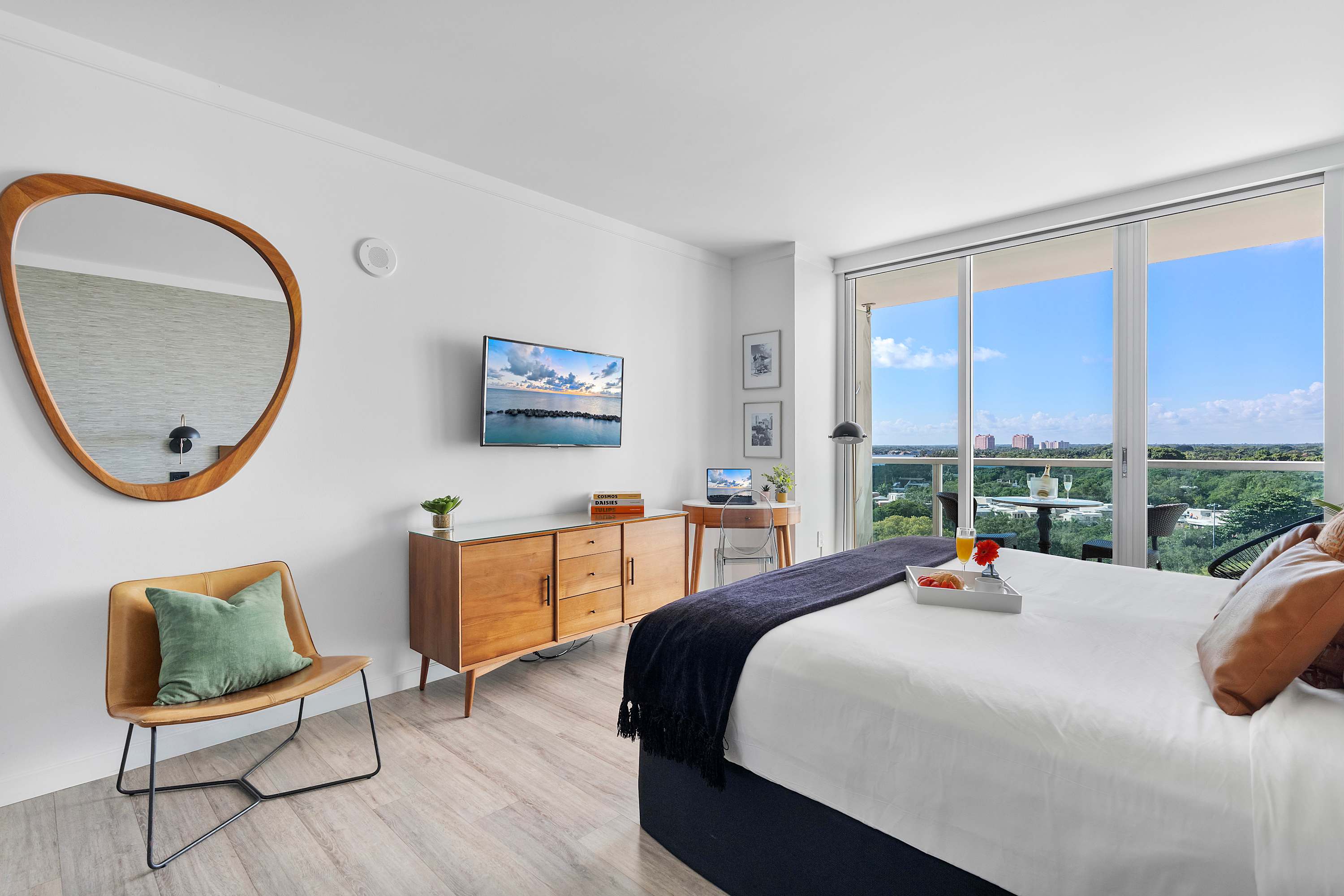 Bright studio space with a queen bed, a wooden credenza with a flat-screen TV, and a small round desk with a laptop, all set against light walls and a large window overlooking the bay.