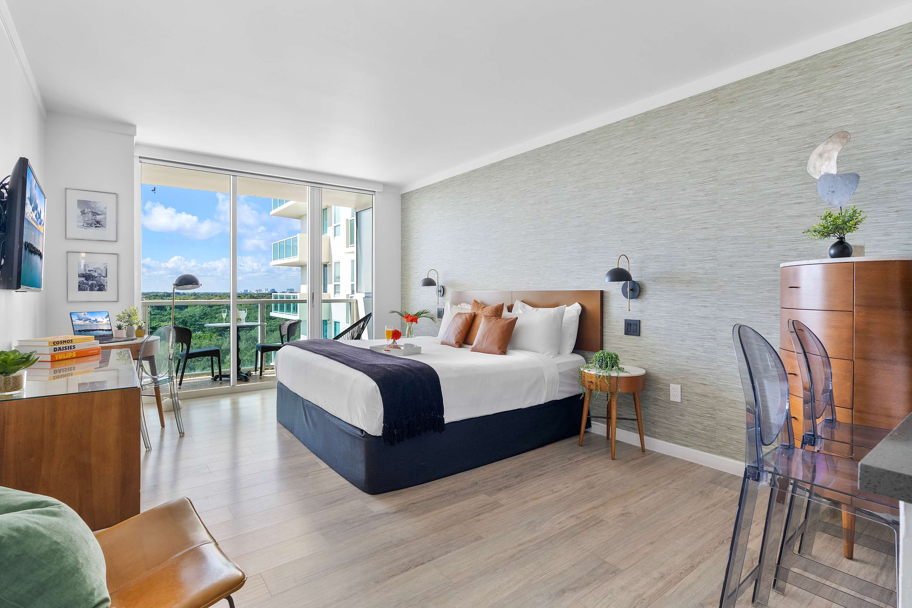 Spacious studio apartment featuring a queen bed with a wooden headboard, a small desk by the window, and a glimpse of the modern kitchen, all with light wood-look flooring and a textured accent wall.