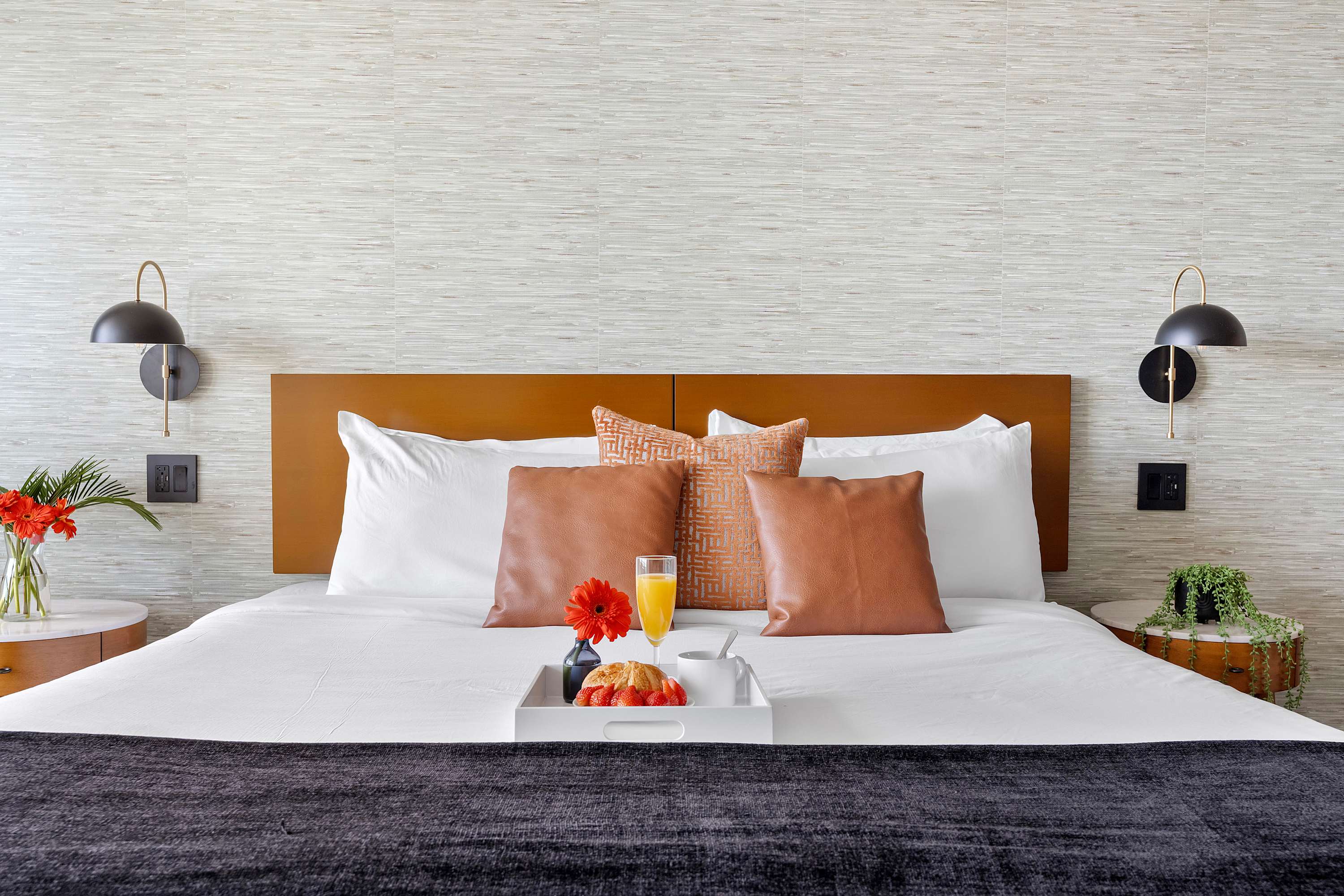 Close-up of a neatly made queen bed with white linens, a dark throw, and decorative orange and brown pillows, flanked by two modern black wall sconces and nightstands against a textured accent wall.