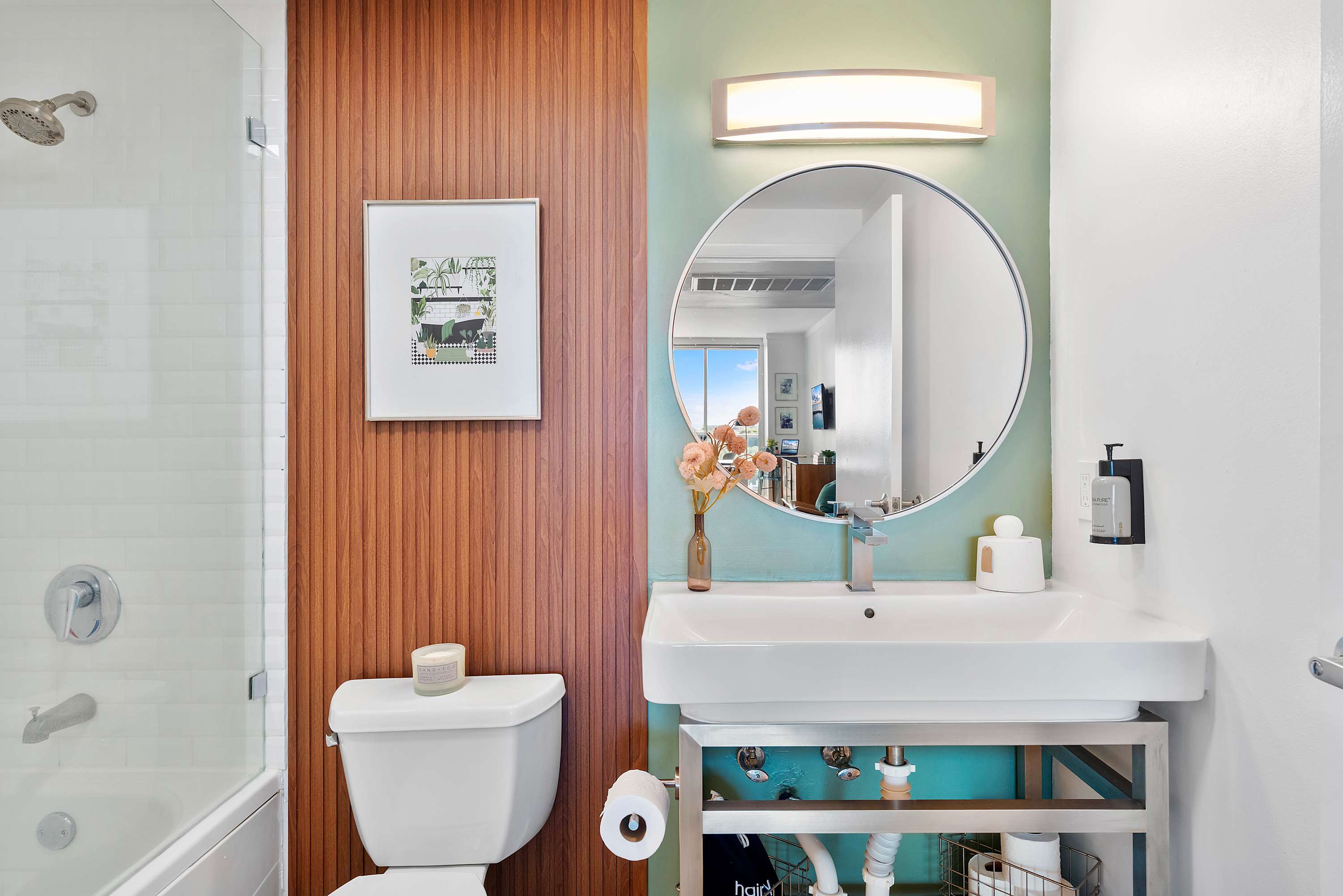 Modern bathroom featuring a white vanity sink with a round mirror, a shower/tub combo, and a striking wooden slatted accent wall contrasting with a light green painted wall.