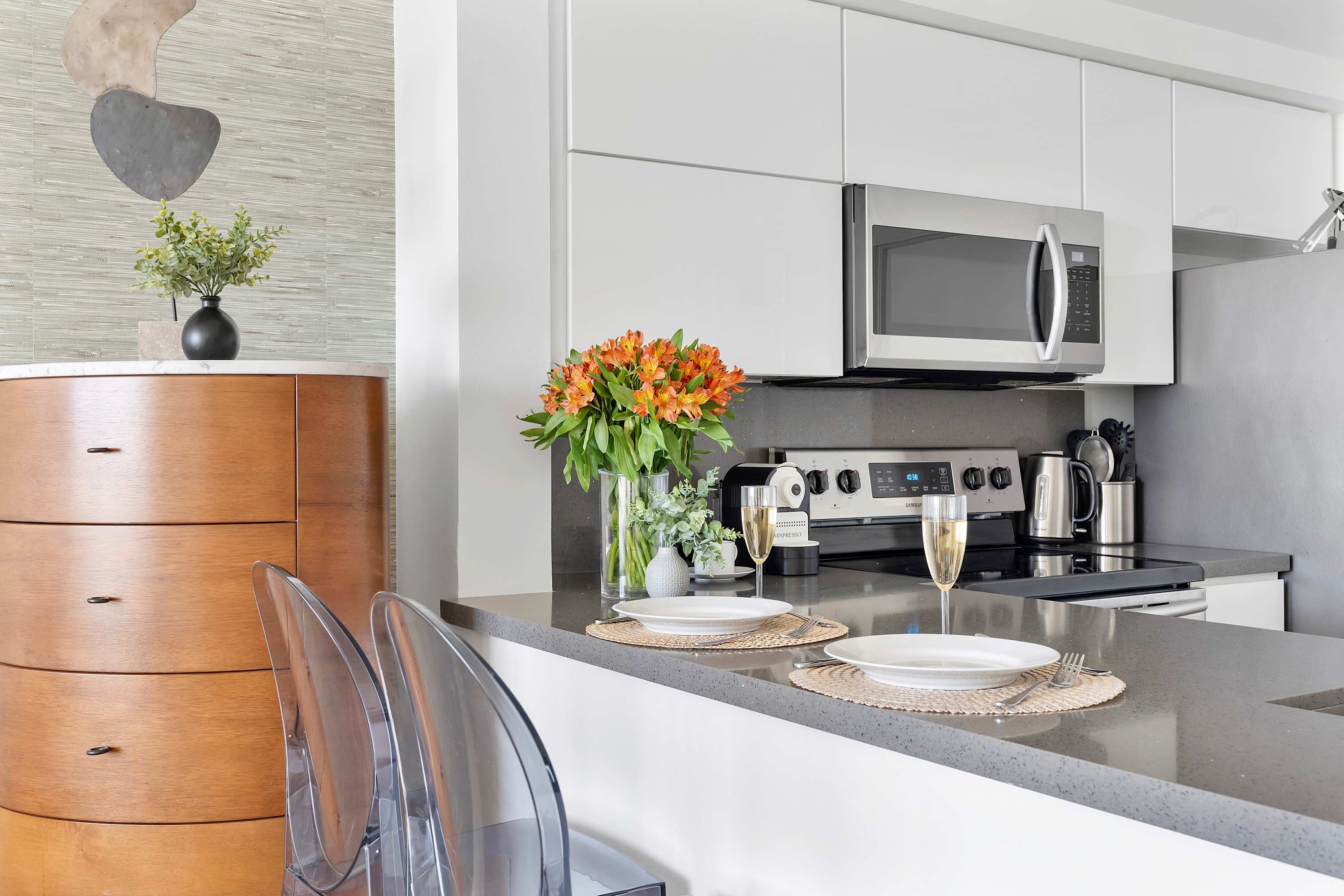 Modern kitchen counter with dark grey countertops, a stainless steel sink, and white flat-panel cabinets, featuring a microwave, stove, and dishwasher, with two clear bar stools.