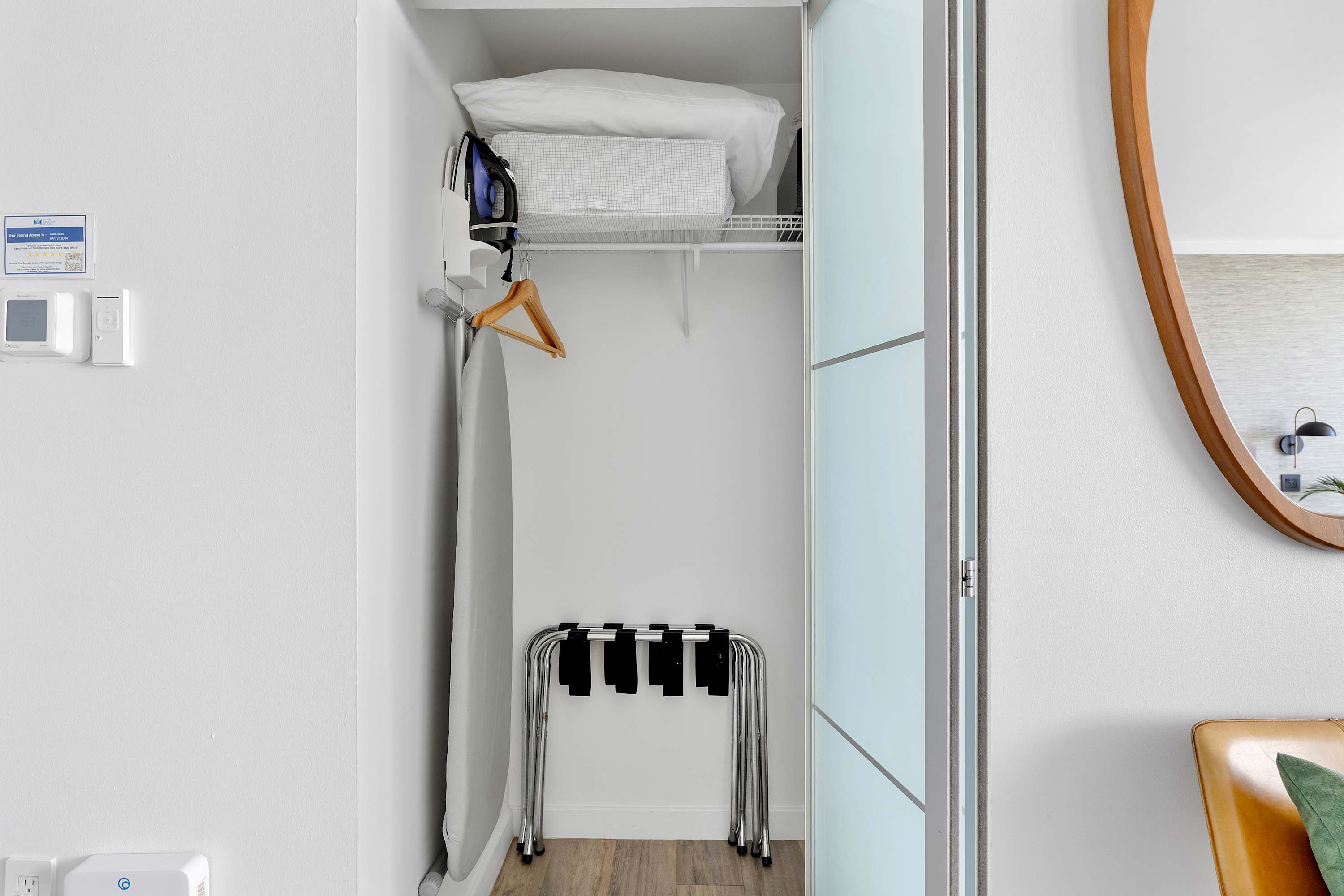 An open closet revealing an ironing board, an iron, a white storage basket, and a metal luggage rack, indicating practical amenities for guests.