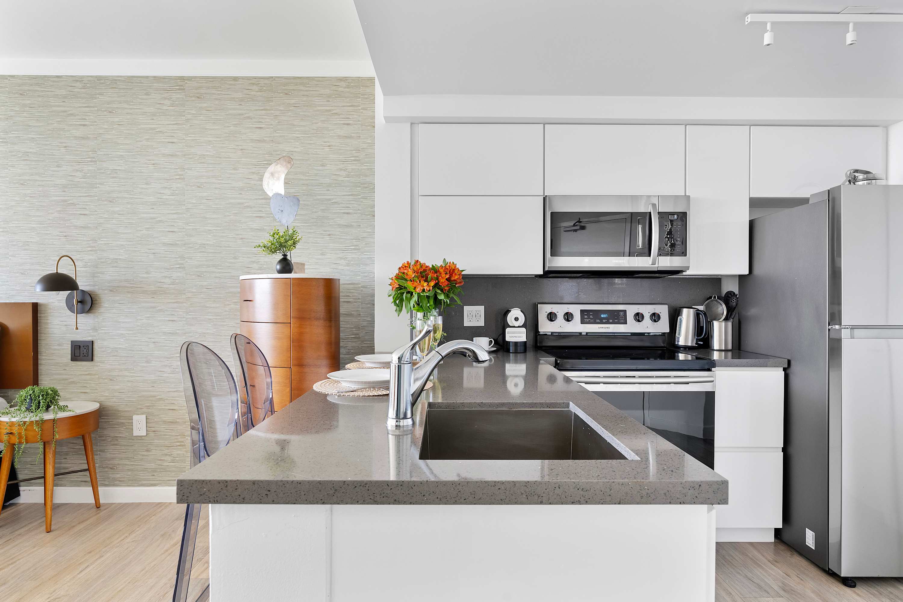 A detailed view of the modern kitchen island with a stainless steel sink and faucet, flanked by two clear acrylic bar stools, and showcasing white cabinetry and stainless steel appliances in the background.
