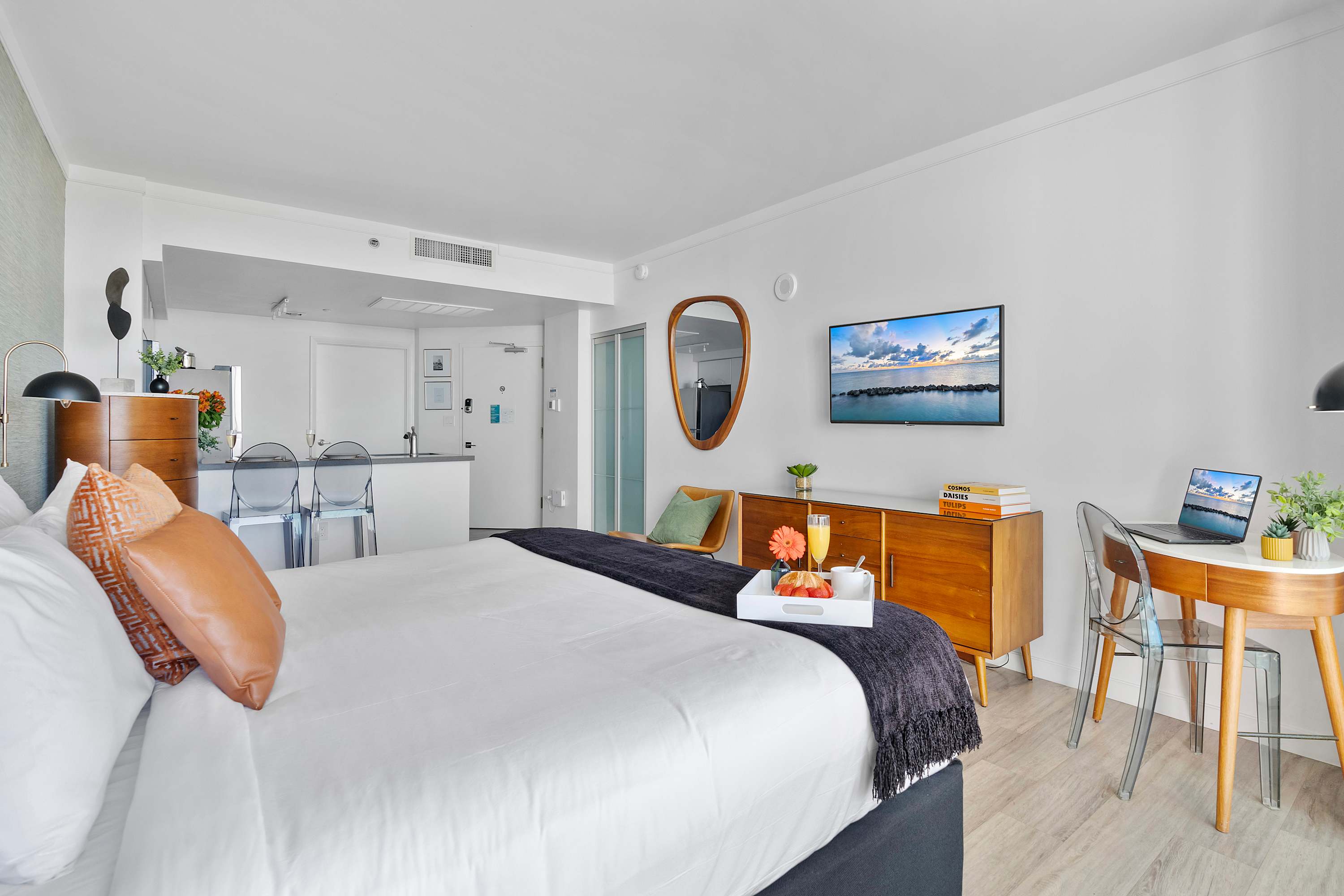 Studio apartment interior showing the queen bed in the foreground, a wooden credenza with a TV, and the modern kitchen area with bar stools in the background, leading to the entrance.