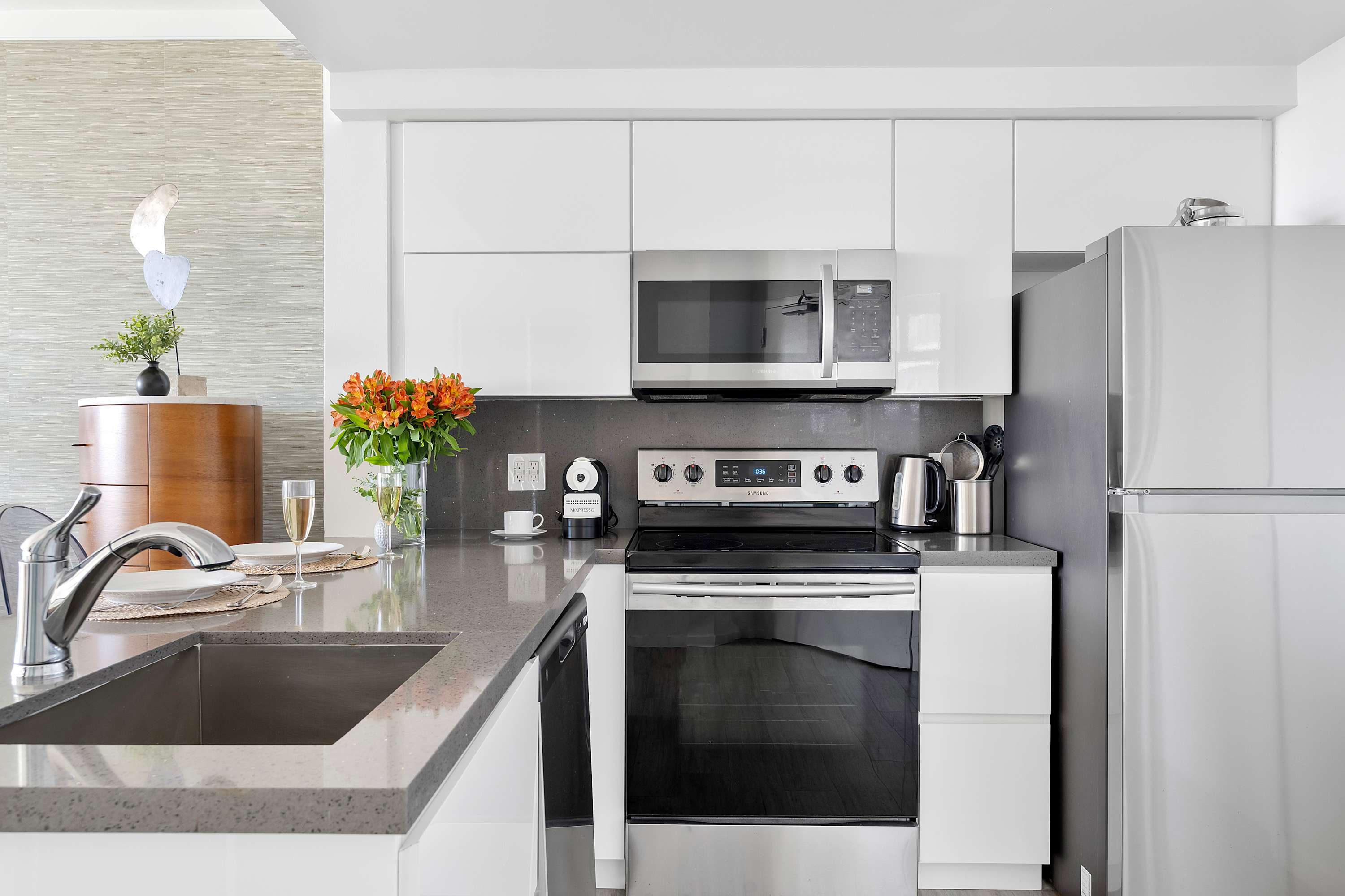 A modern kitchen featuring sleek white cabinetry, stainless steel appliances including a microwave, oven, and refrigerator, and a dark grey countertop with an undermount sink. A vase of orange flowers adds a pop of color.