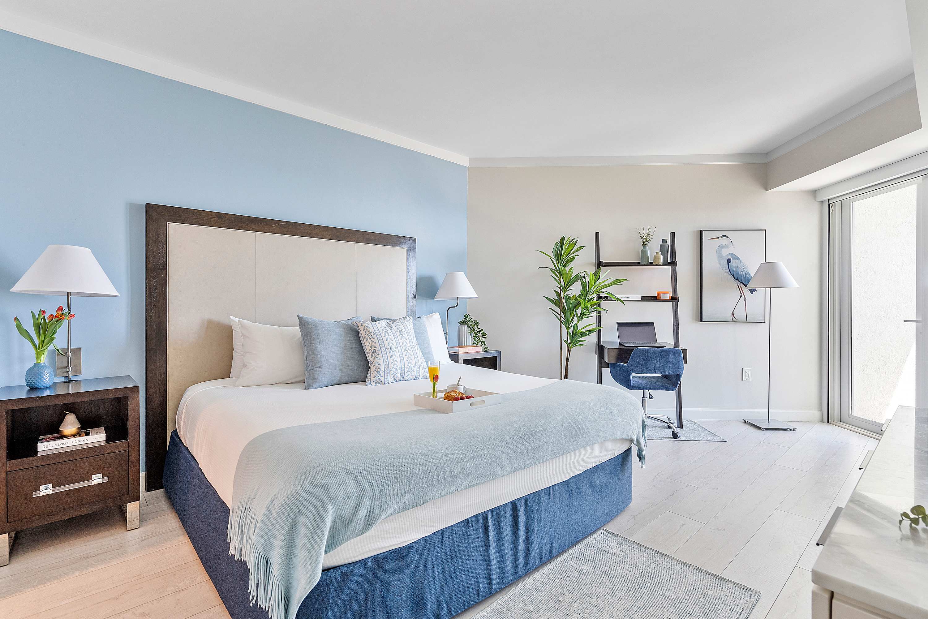 A bright bedroom with a king-sized bed featuring a tall upholstered headboard, a light blue accent wall, and light wood flooring. A dark wood nightstand with a lamp and flowers sits beside the bed, and a small desk area with a plant is visible on the right.