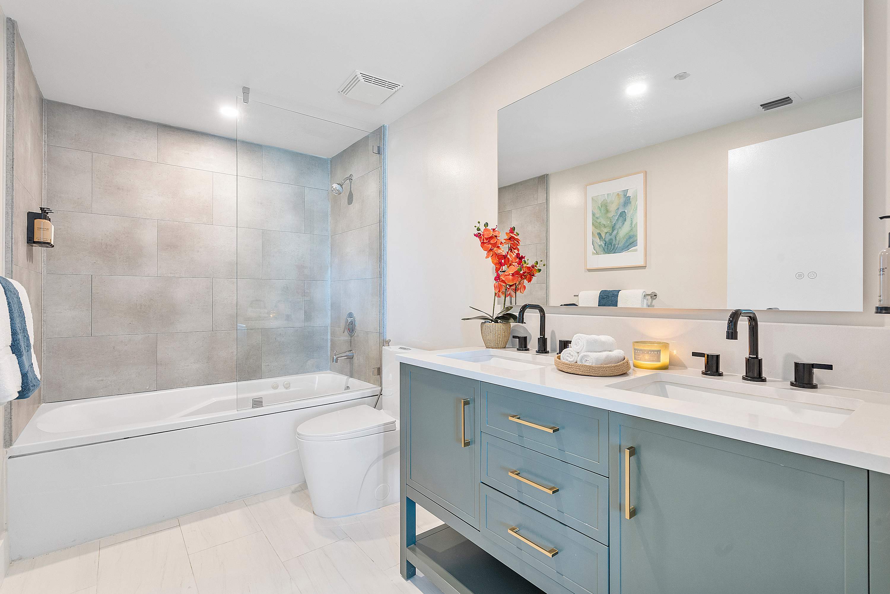 A modern bathroom with a light gray tiled shower/tub combo, a white toilet, and a double vanity with a teal cabinet and white countertop. Black fixtures and a large mirror complete the space, with orange flowers adding a pop of color.