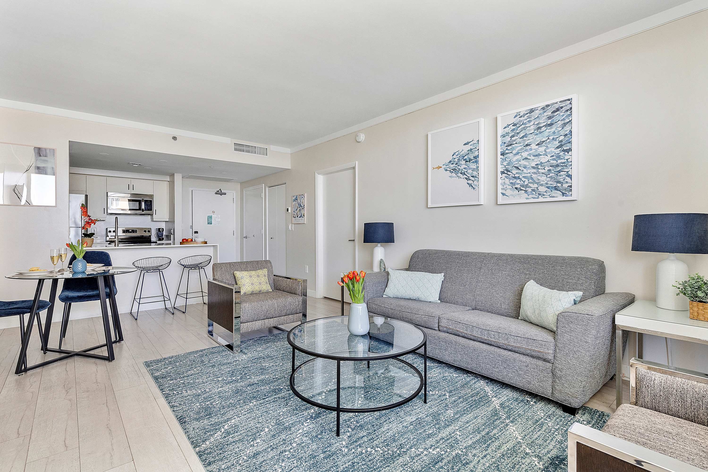Open-concept living area with a gray sofa, a gray armchair, a glass coffee table, and a blue rug, leading into a compact kitchen with white cabinets and stainless steel appliances.