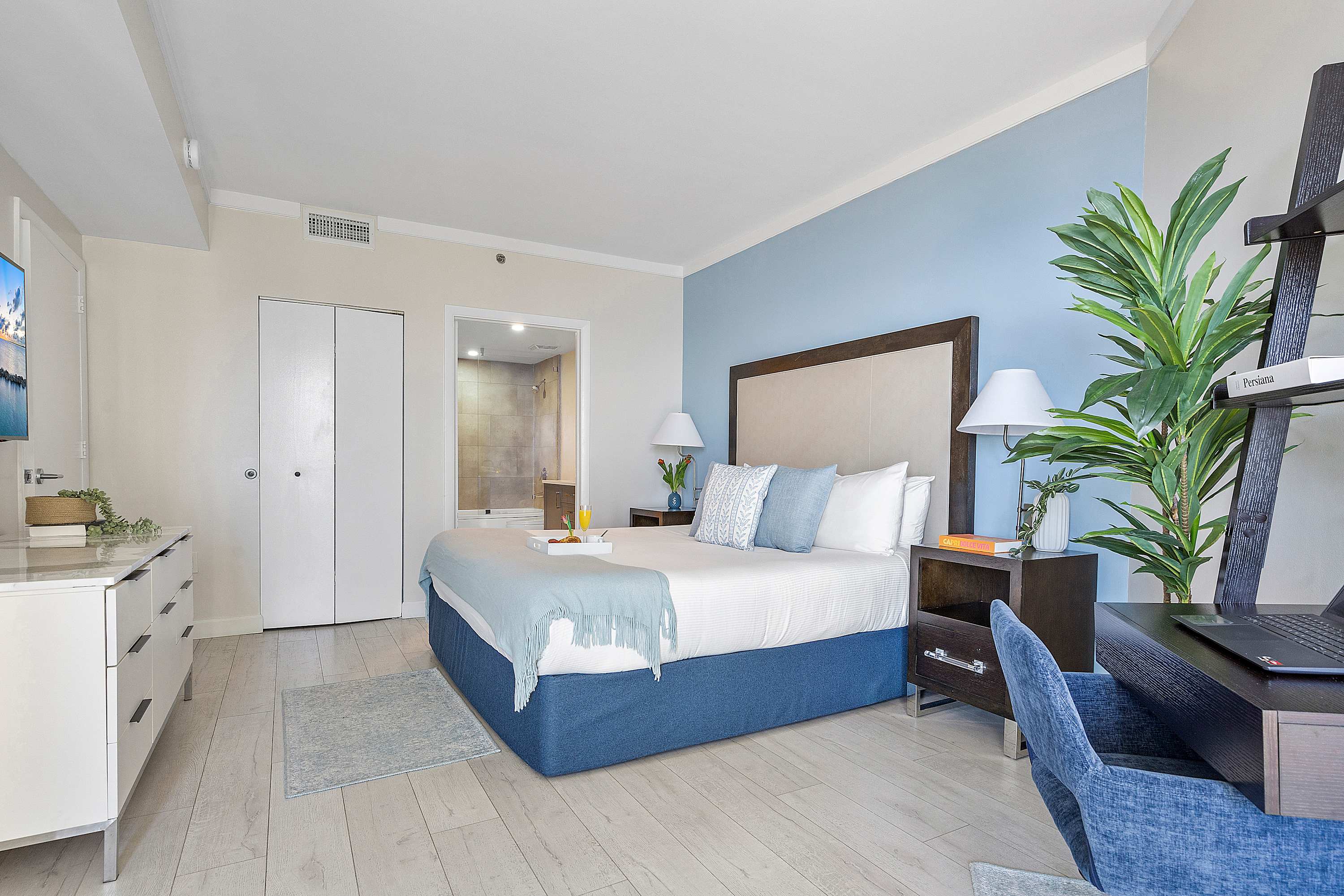 A wide shot of the bedroom showing the king-sized bed with a blue bed skirt and light blue throw, a white dresser, and a dark wood desk with a blue chair. A large plant stands next to the desk, and a doorway leads to the bathroom.