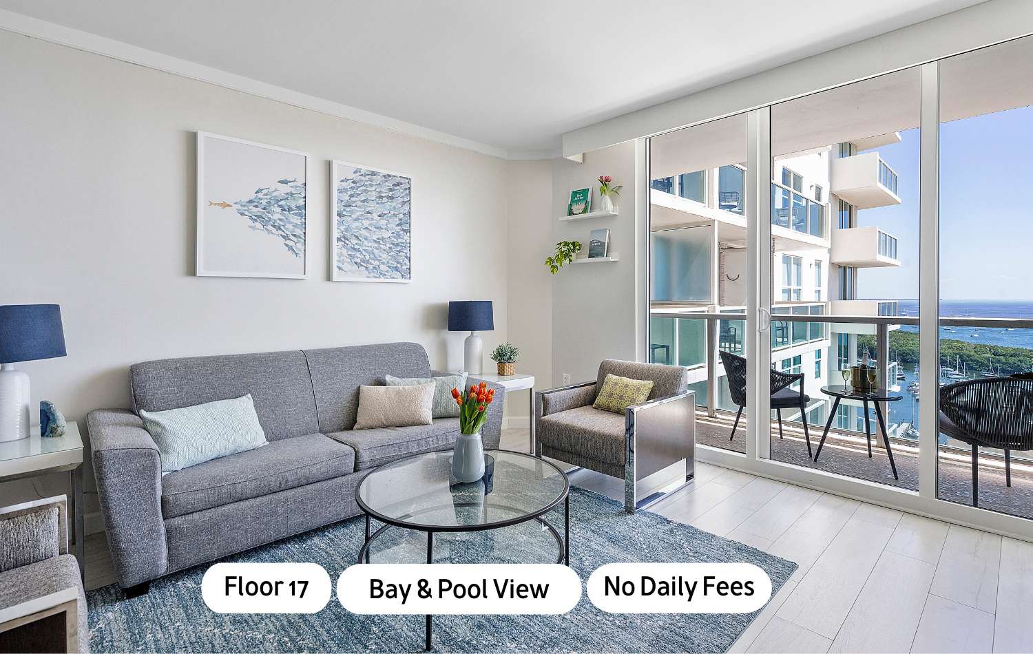 Bright living room with a gray sofa, a gray armchair, a glass coffee table, and a blue patterned rug, featuring large sliding glass doors leading to a balcony with bay and city views.