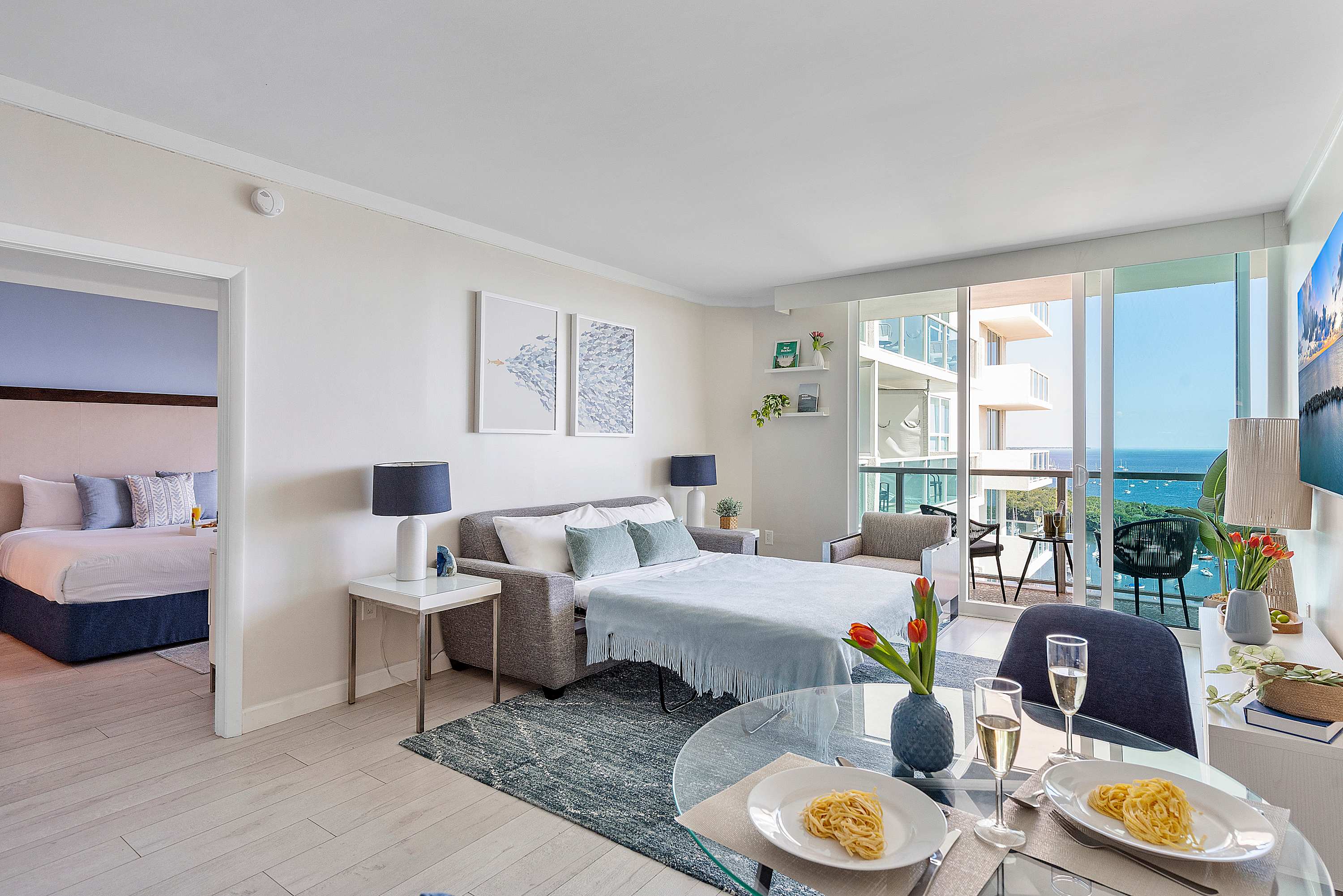An open-concept living and dining area with a pull-out sofa bed, a glass dining table set for two, and light wood flooring. Large sliding glass doors lead to a balcony with ocean views.