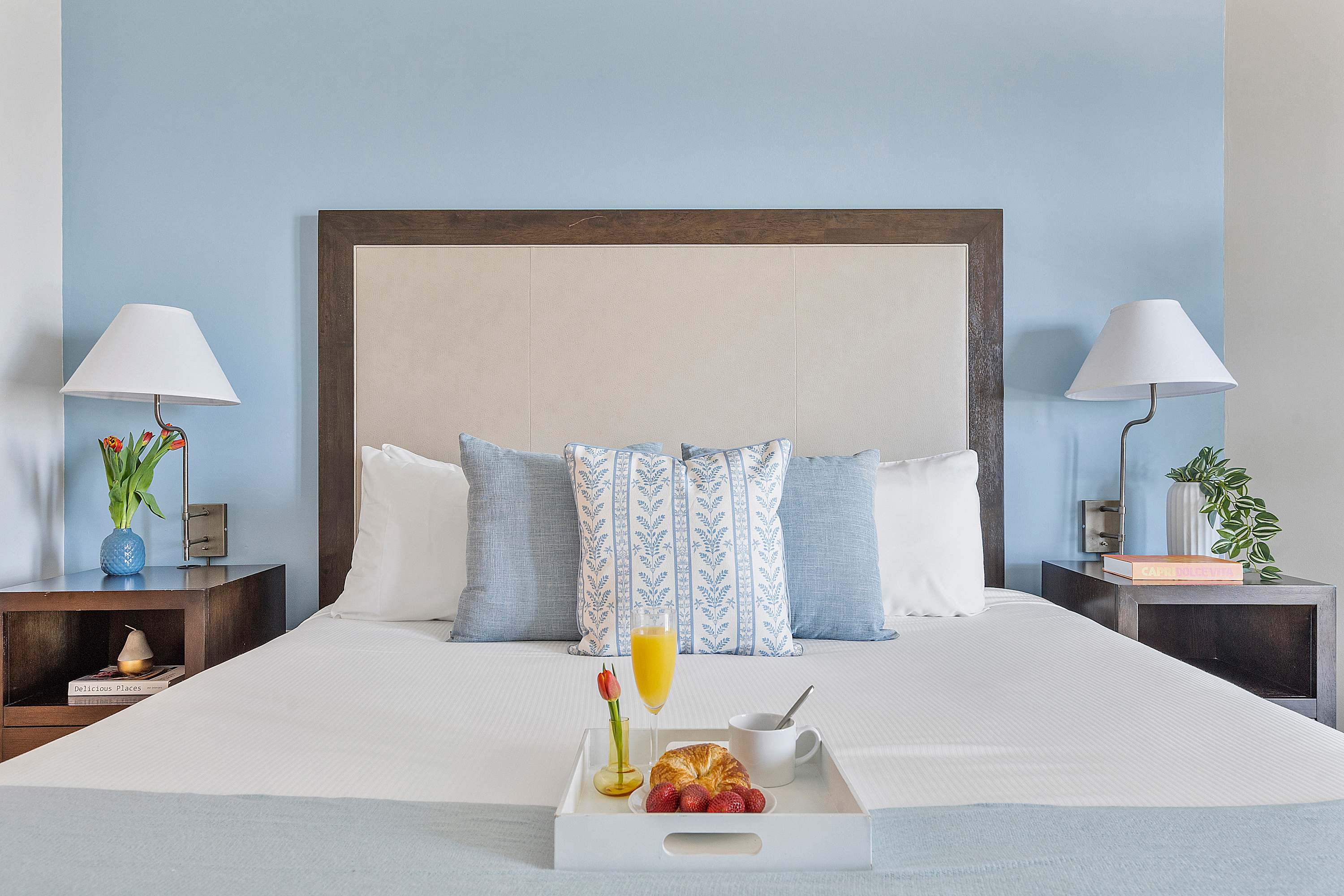 Serene bedroom featuring a large bed with a light blue headboard, white bedding, and blue patterned pillows, flanked by dark wood nightstands with lamps and a breakfast tray.