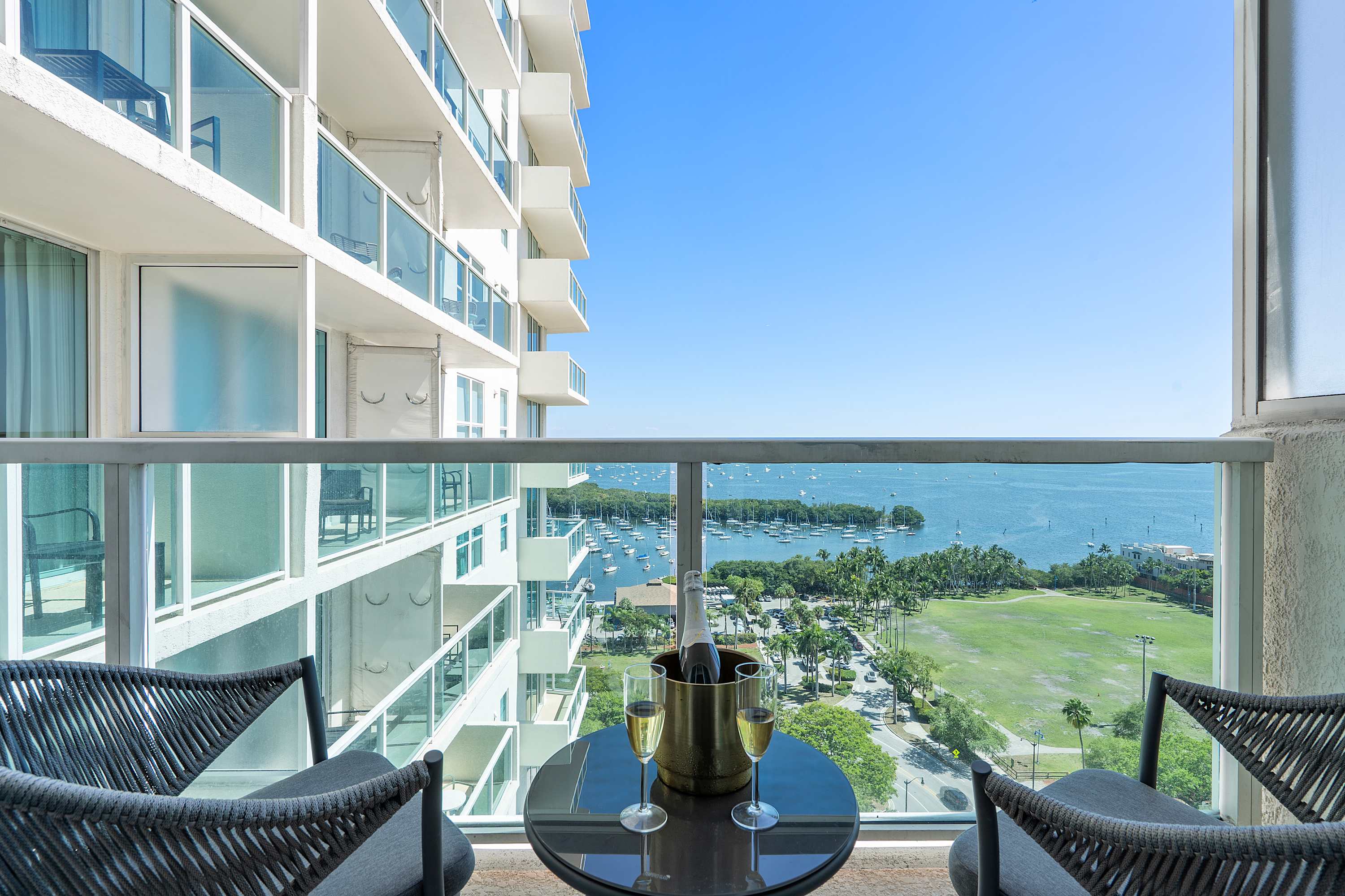 A private balcony with two woven chairs and a small round table, offering panoramic views of a bay filled with boats and a distant city skyline. A bottle of champagne and two flutes are on the table.