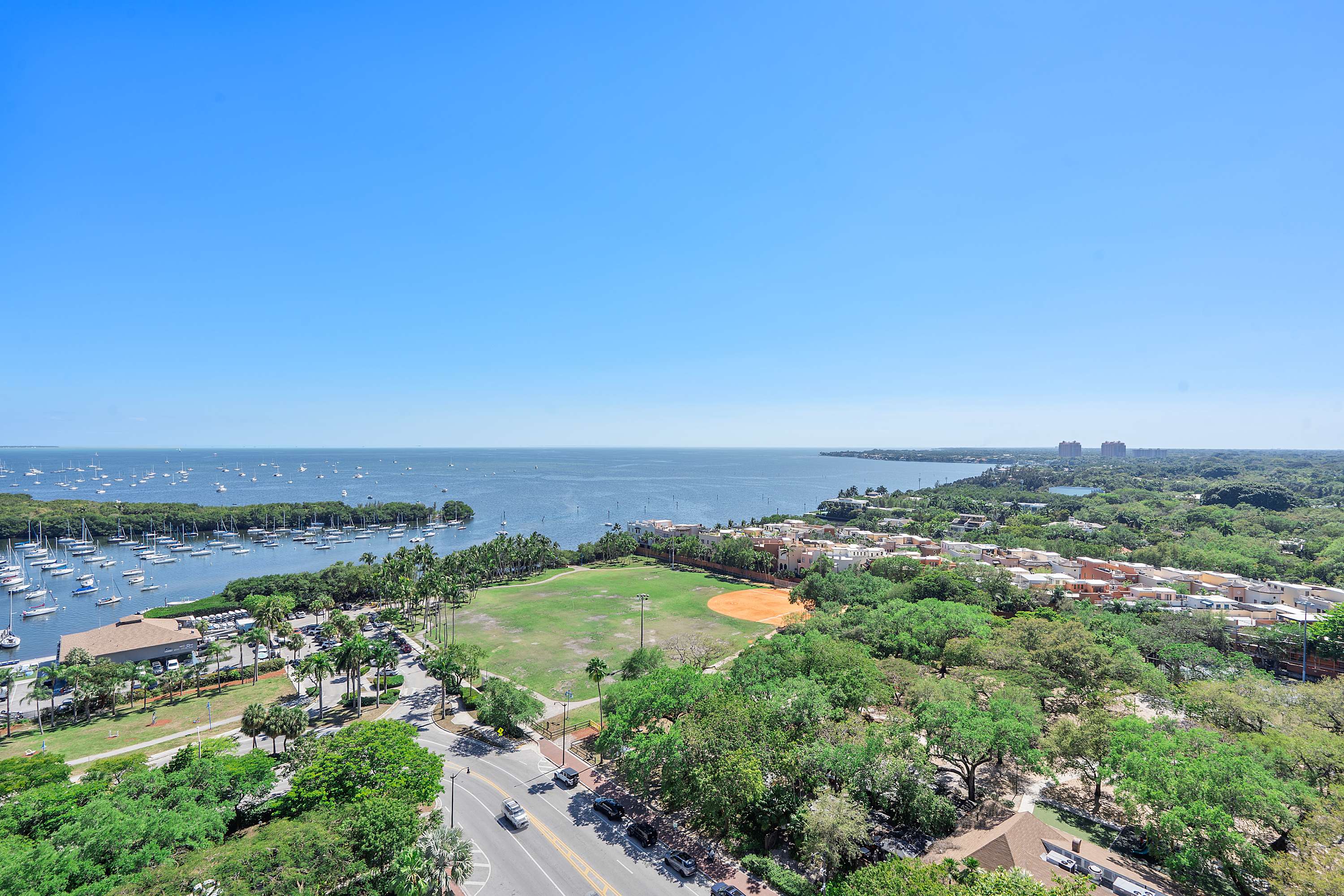 High-angle aerial view showcasing a vast blue bay filled with numerous boats, a green baseball field, and a residential area surrounded by lush trees under a clear blue sky.