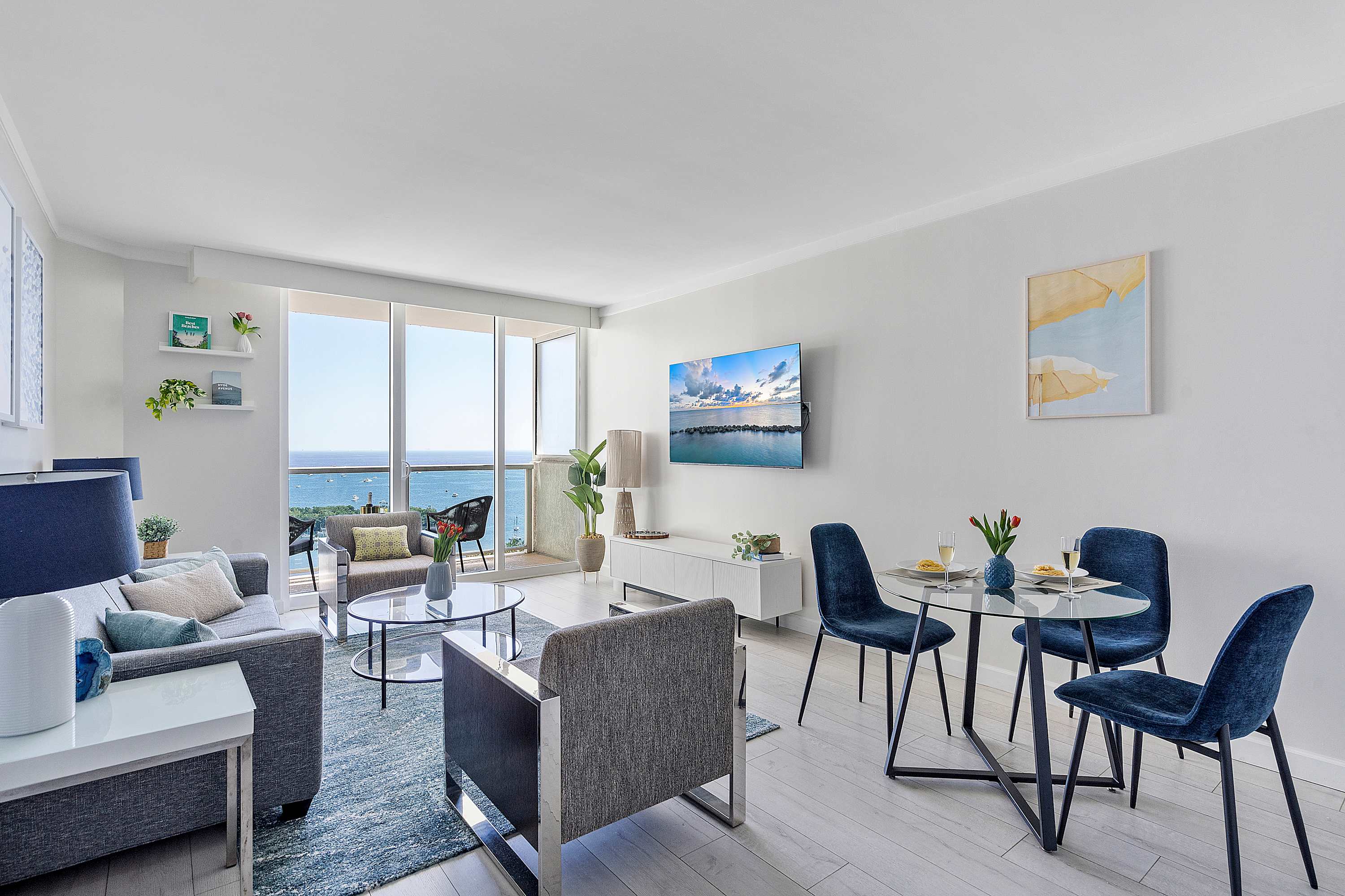 Modern living room with a gray sofa, a gray armchair, a glass coffee table, and a blue rug, featuring a wall-mounted TV and large windows offering ocean views. A small dining area is visible to the right.