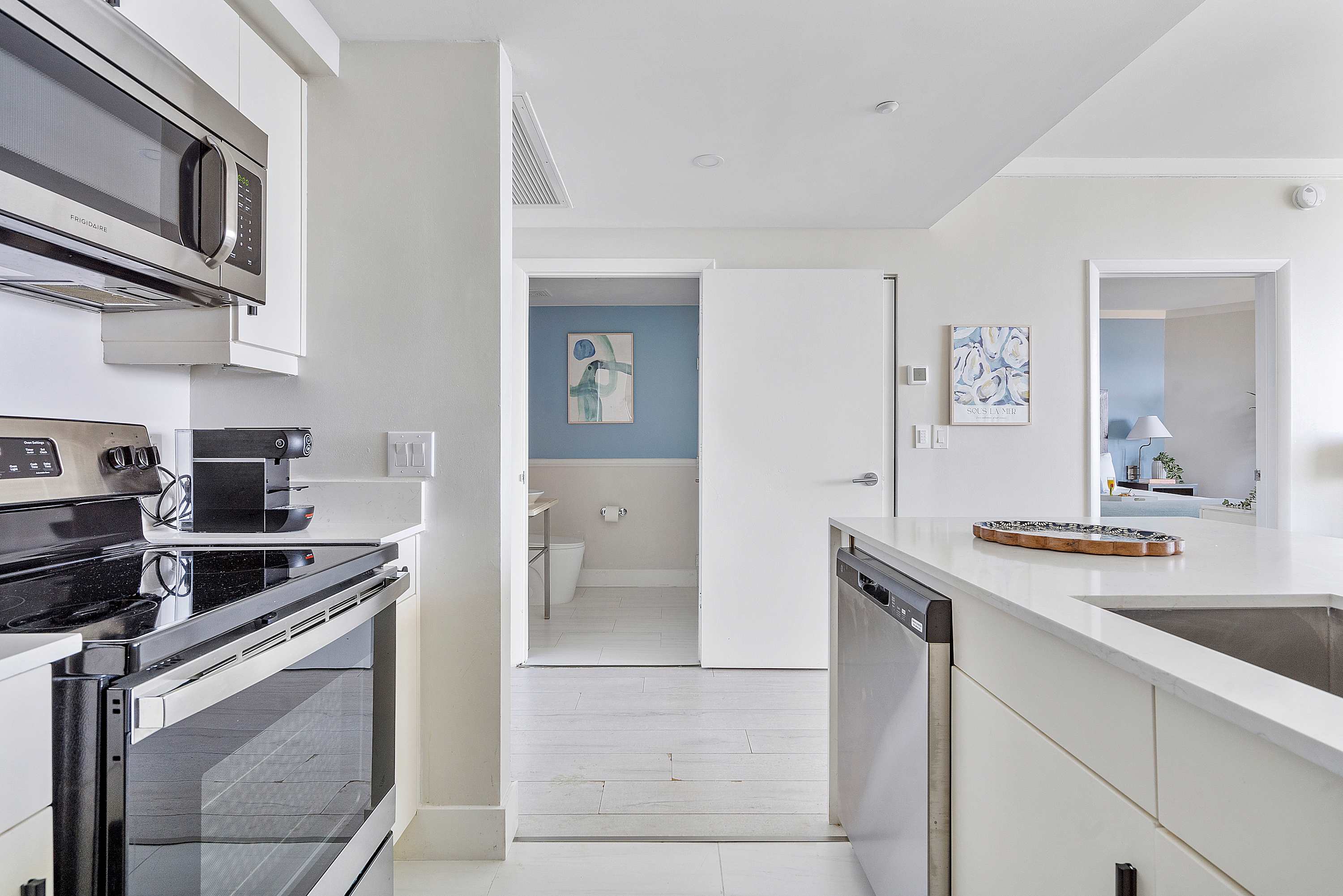 View from the kitchen showing a white countertop island with a sink, leading towards a light blue half bathroom with a vessel sink and a round illuminated mirror.