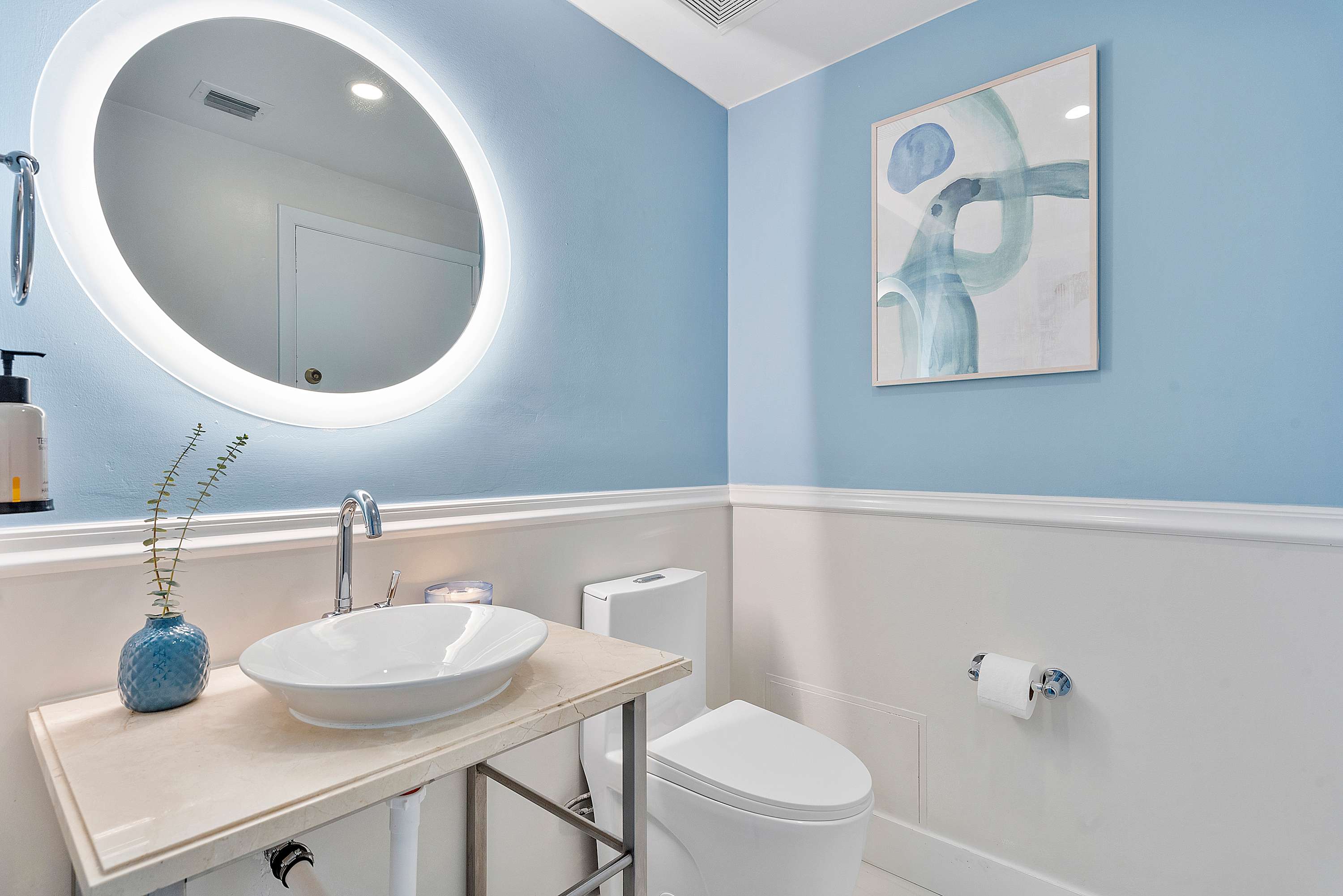 Modern half bathroom with light blue walls and white wainscoting, featuring a white vessel sink on a light-colored vanity, a toilet, and a large round illuminated mirror.