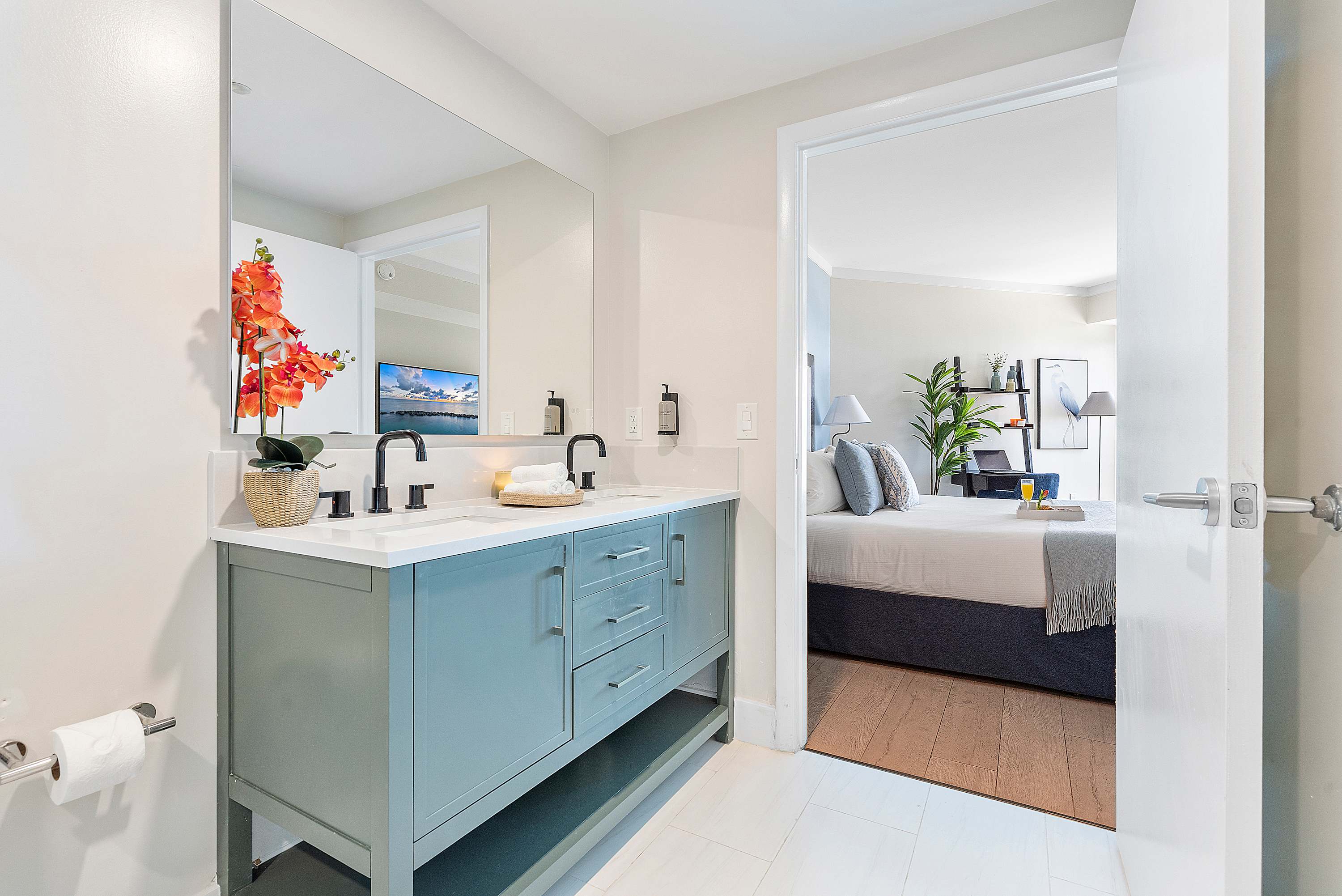 A view of the double vanity in the modern bathroom, featuring a teal cabinet, white countertop, and black fixtures. A large mirror reflects the bedroom, and vibrant orange flowers add a decorative touch.