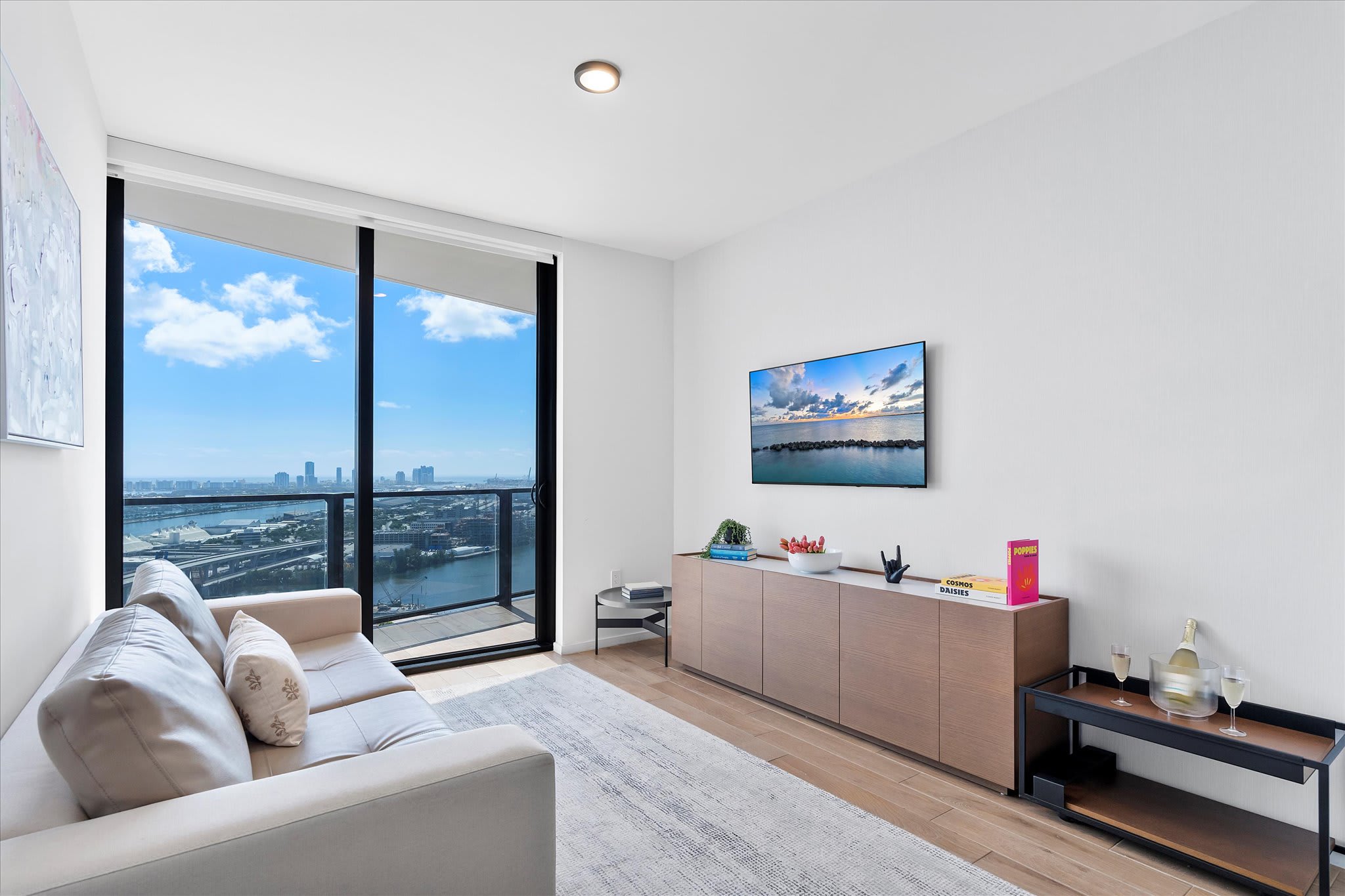 A bright living room with a light-colored sofa, a wall-mounted TV, and a long wooden console table, featuring a large sliding glass door that opens to a balcony with panoramic city and water views.