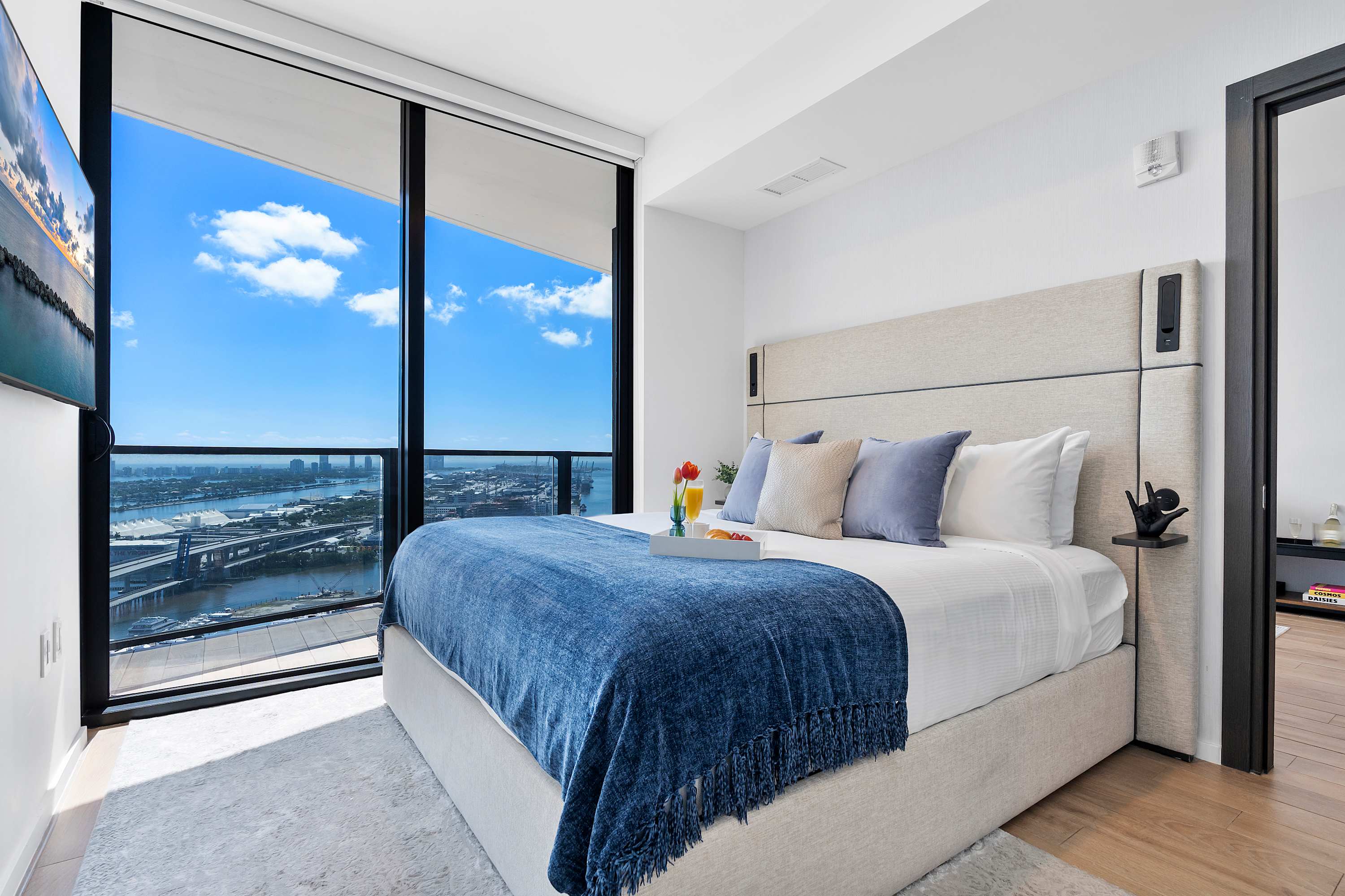A serene bedroom featuring a large bed with a beige headboard and blue accents, positioned to enjoy expansive city and water views through a floor-to-ceiling window.