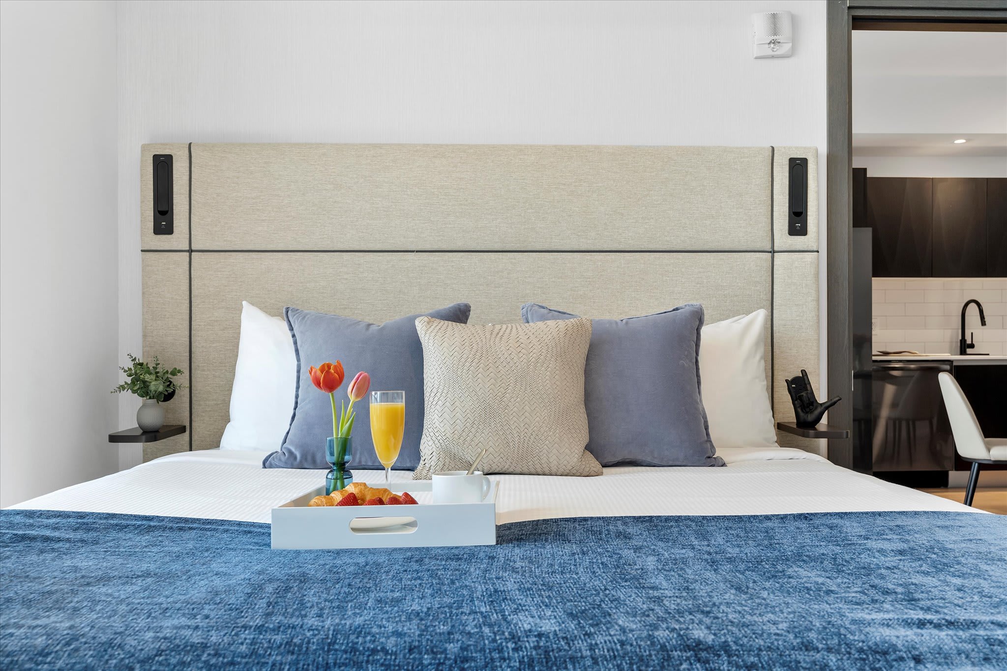 A wide shot of a modern bedroom with a large bed, upholstered headboard, and a mix of blue and beige pillows. A breakfast tray with flowers and food is centered on the bed, with a partial view of a kitchen in the background.
