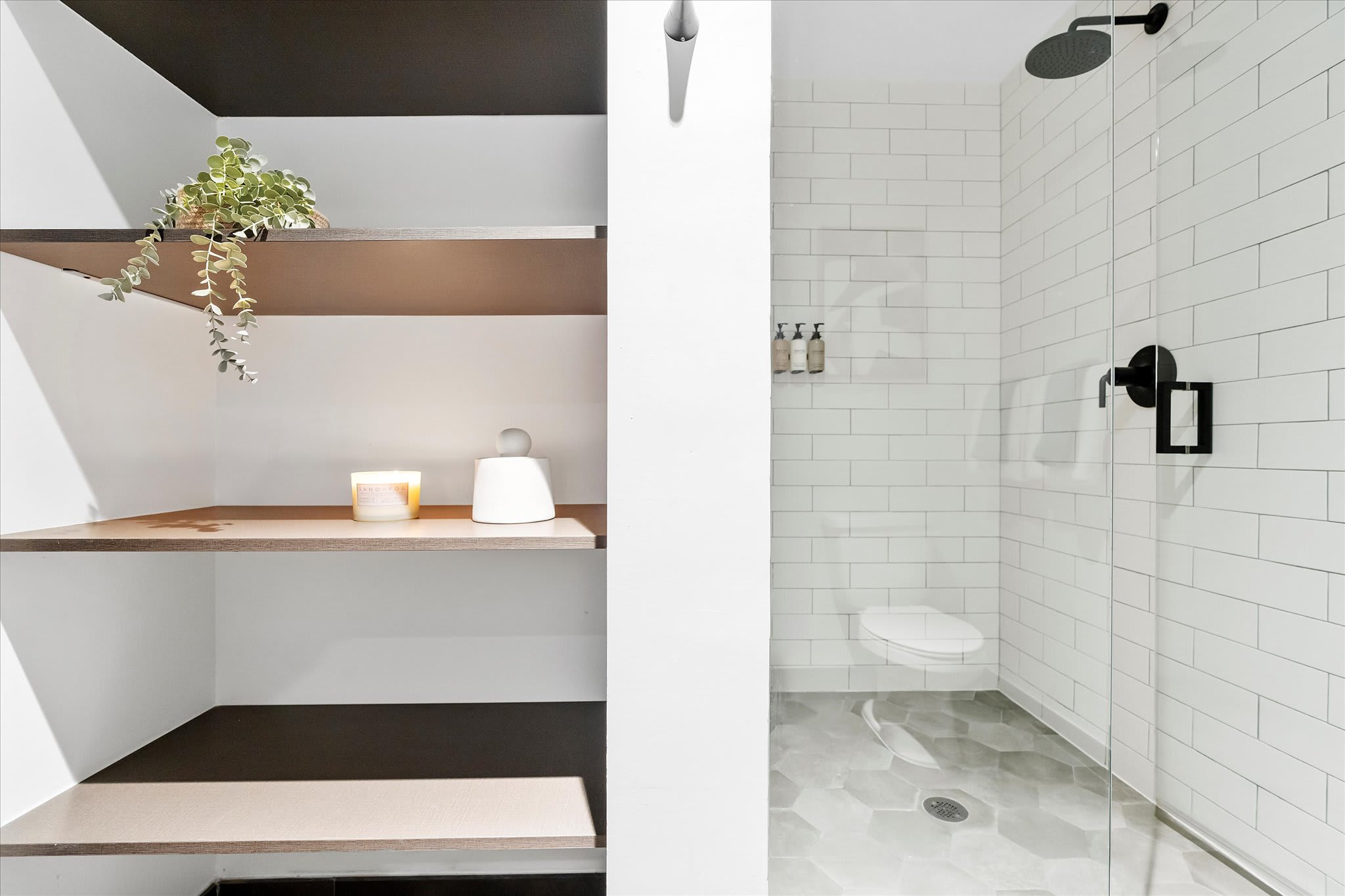 A contemporary bathroom showcasing a walk-in shower with white subway tiles and a black rain shower head, alongside open shelving decorated with a plant and a candle.