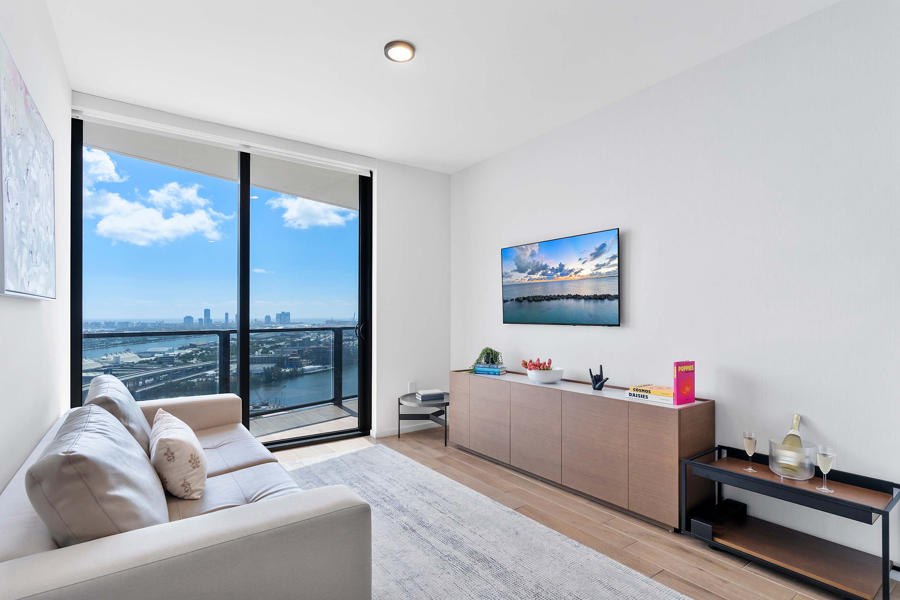 A bright living room with a light-colored sofa, a wall-mounted TV, and a long wooden console table, featuring a large sliding glass door that opens to a balcony with panoramic city and water views.