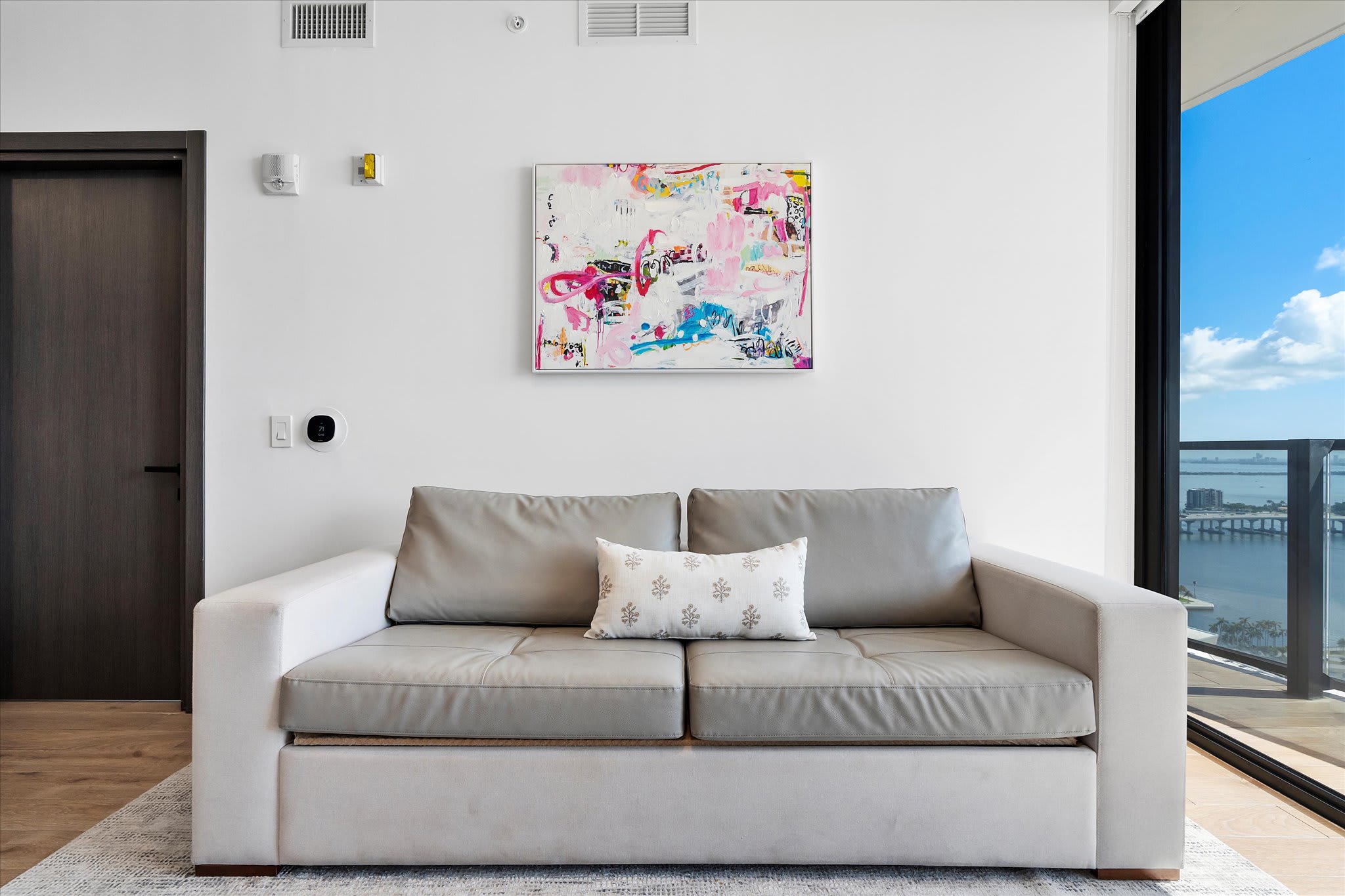 A minimalist living room featuring a light grey sofa bed, abstract artwork on the wall, and a large window offering expansive views of the city and water.