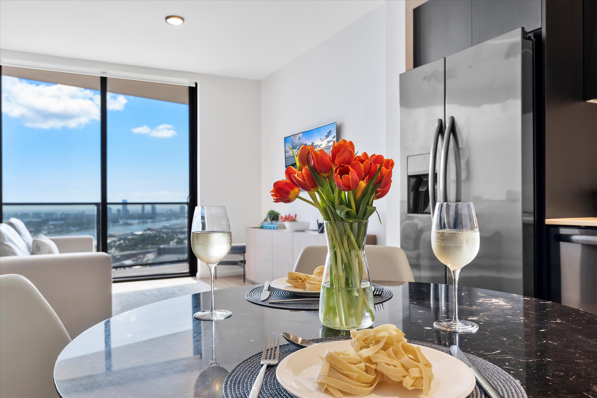 A stylish dining area featuring a round black marble table set with two places, including plates of pasta and wine glasses, centered by a vibrant vase of orange tulips. A modern kitchen with stainless steel appliances is visible in the background, and large windows offer city views.