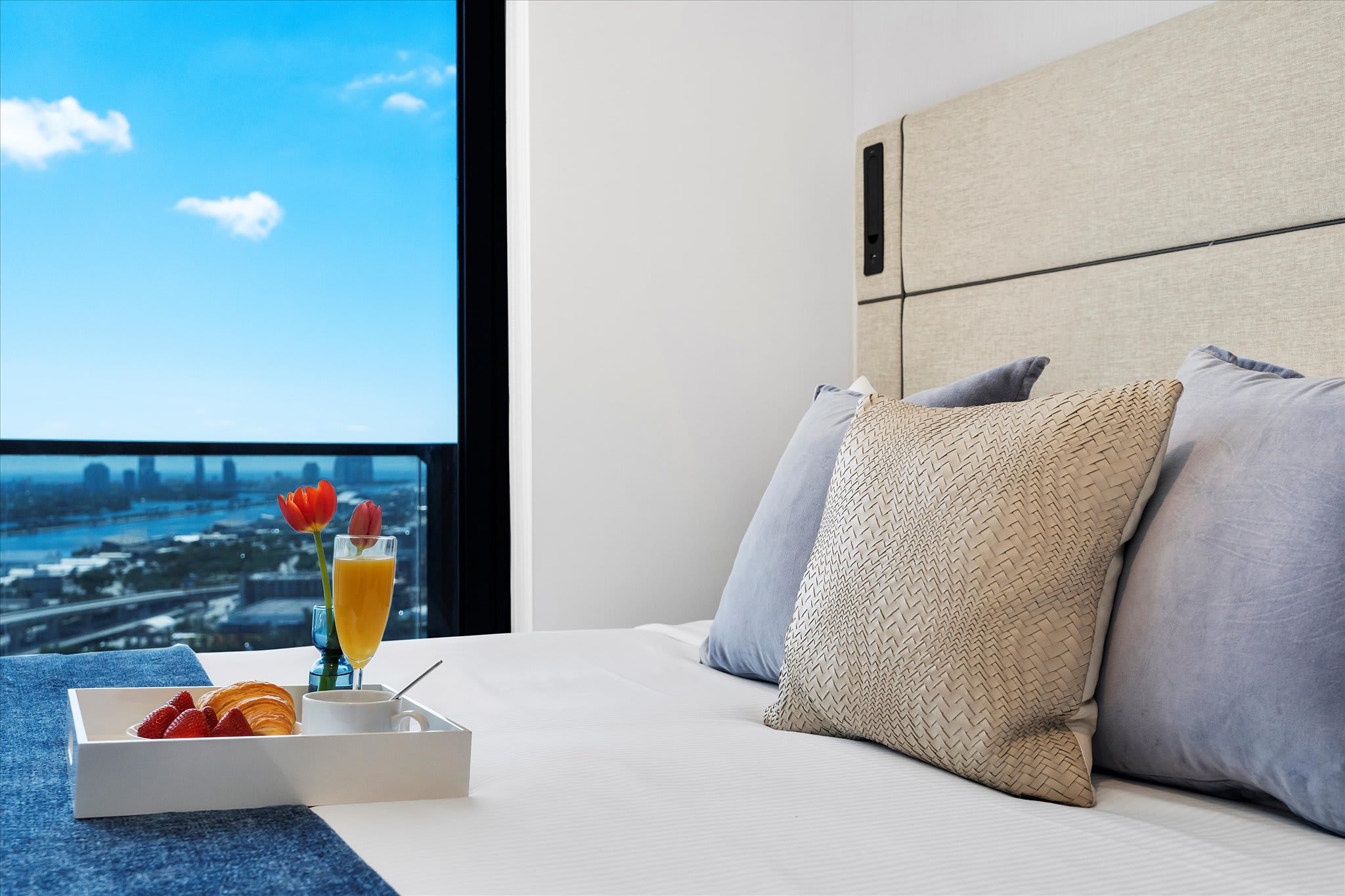 A cozy bedroom scene featuring a breakfast tray with fresh fruit, croissant, orange juice, and coffee on a neatly made bed, with a stunning city and water view visible through a large window.