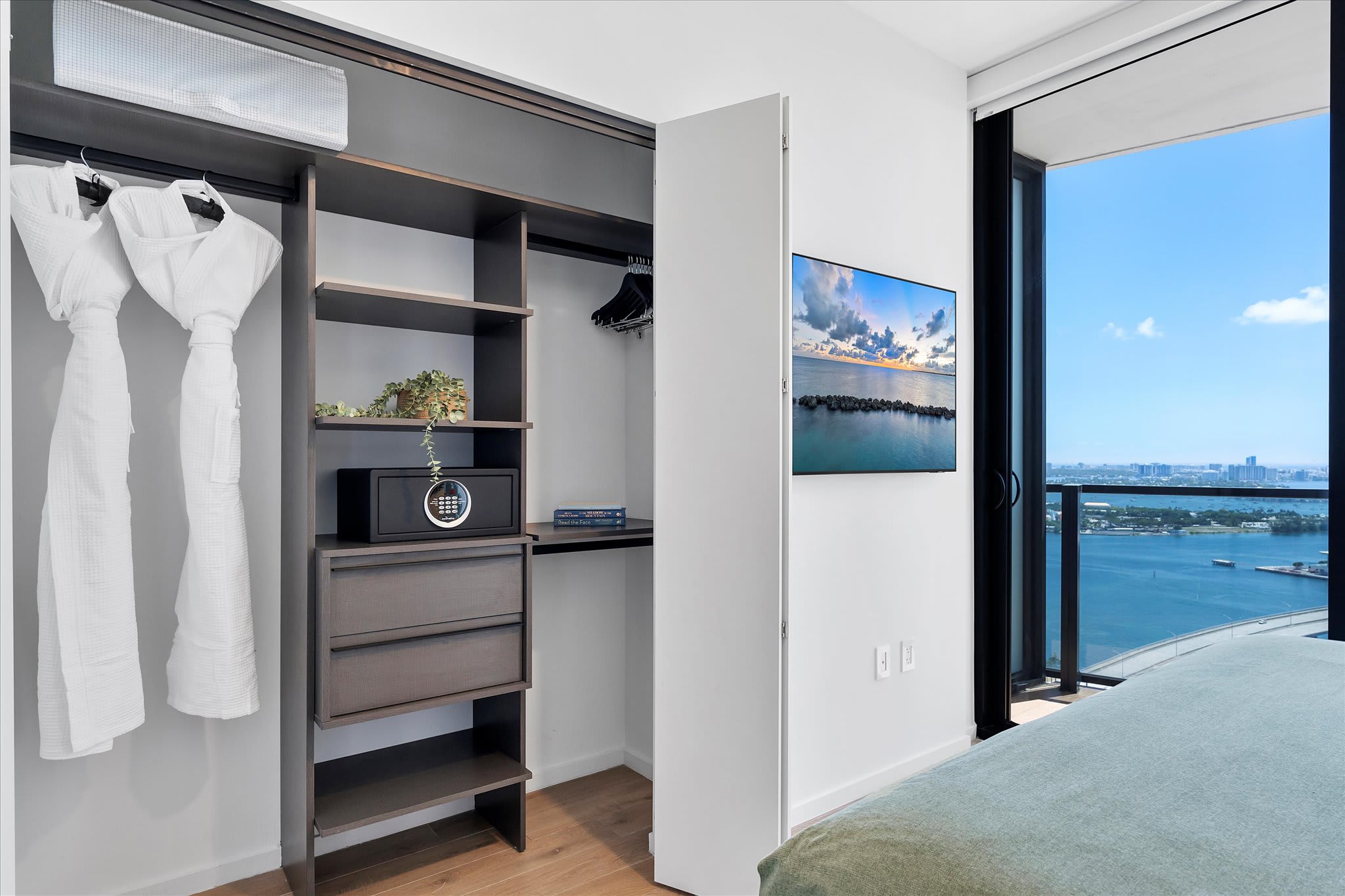 A contemporary bedroom with a wall-mounted television displaying a scenic view, a large window offering expansive city and water views, and an open closet with shelving and hanging space.