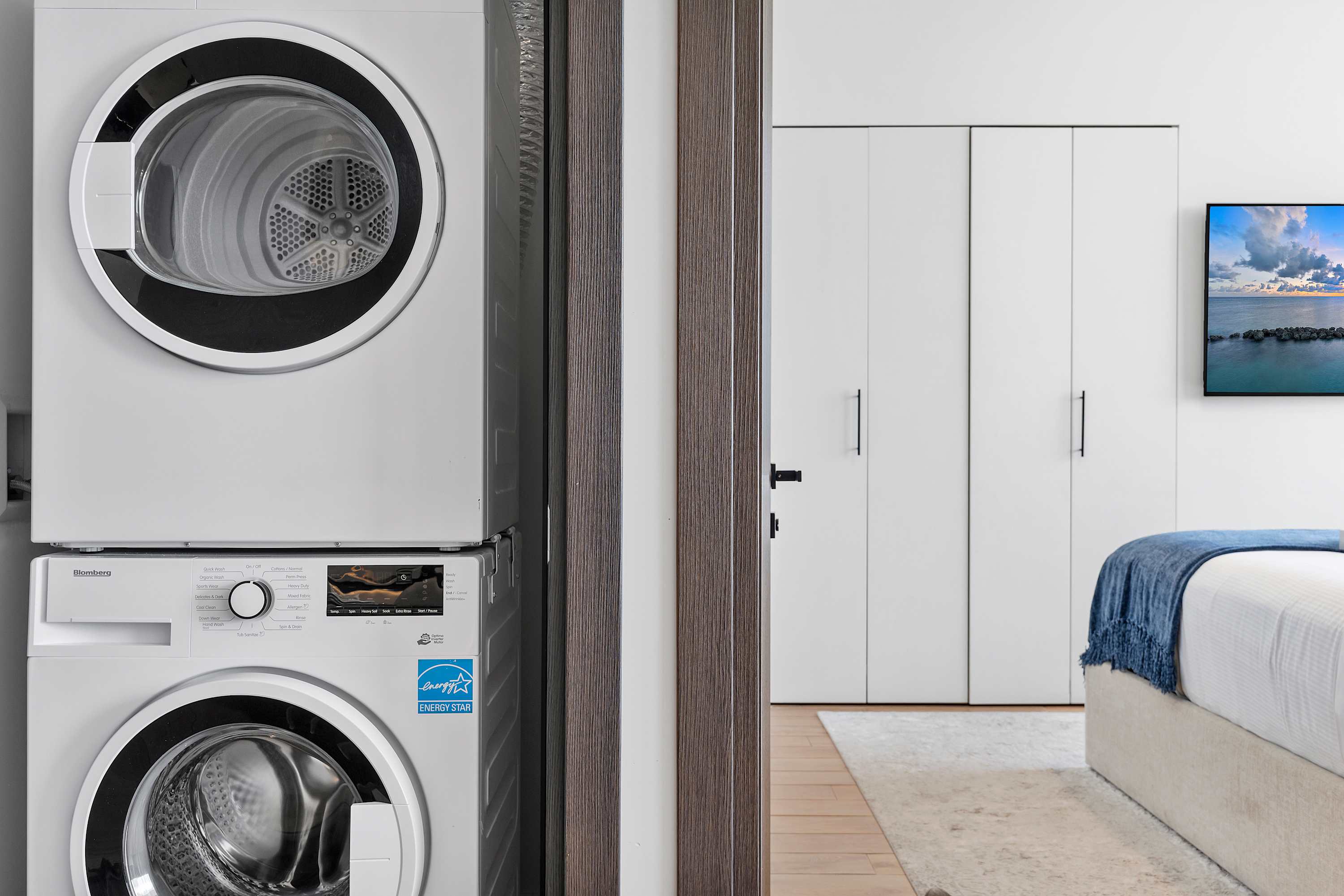 A compact laundry area featuring a stacked, energy-efficient washer and dryer unit, positioned near a doorway that offers a view into the adjacent bedroom with its bed and wall-mounted TV.