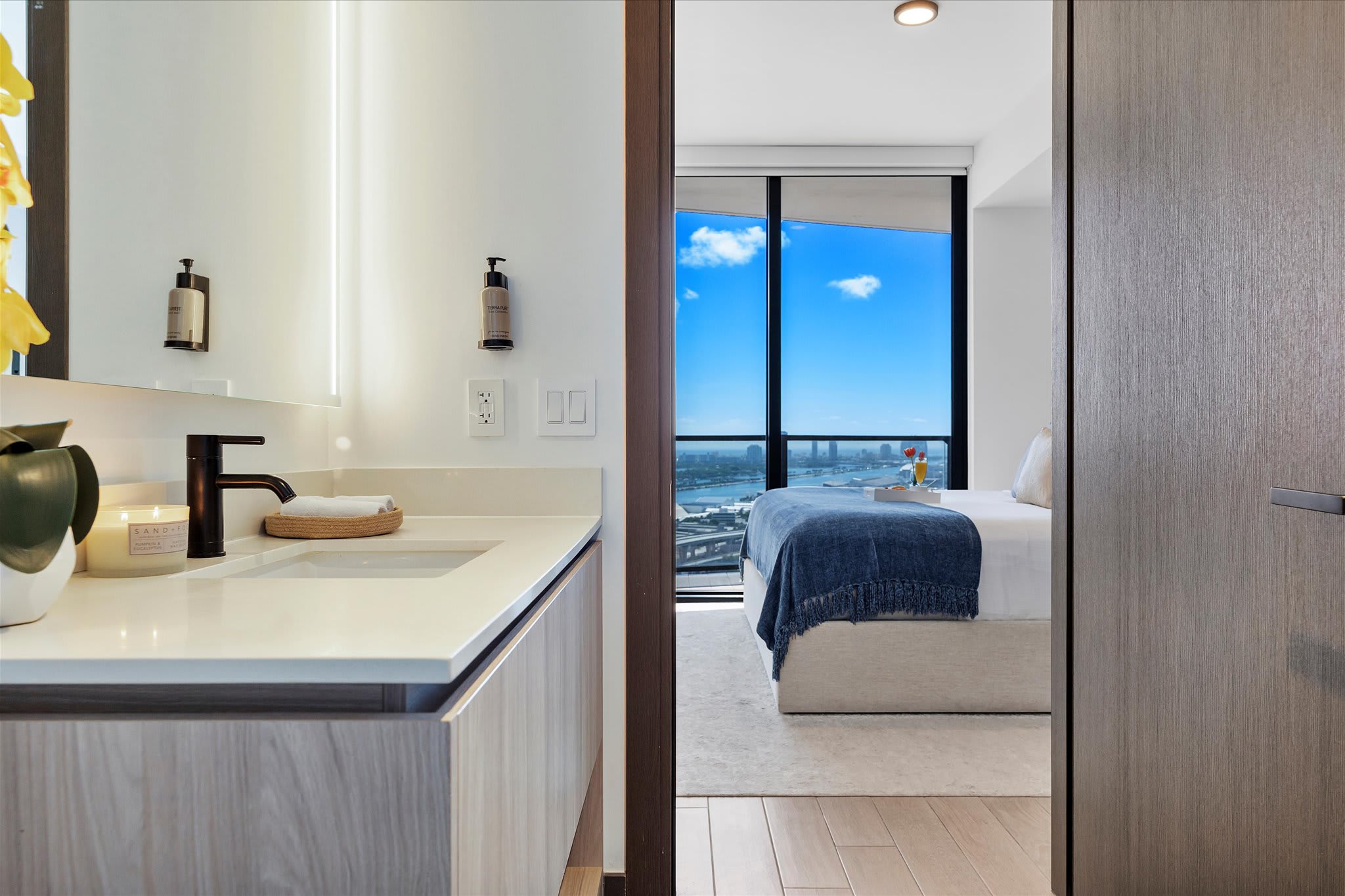 A view from the bathroom entrance, showcasing a modern vanity with a white countertop and sink, a large mirror, and a glimpse into the adjacent bedroom with its bed and city view window.