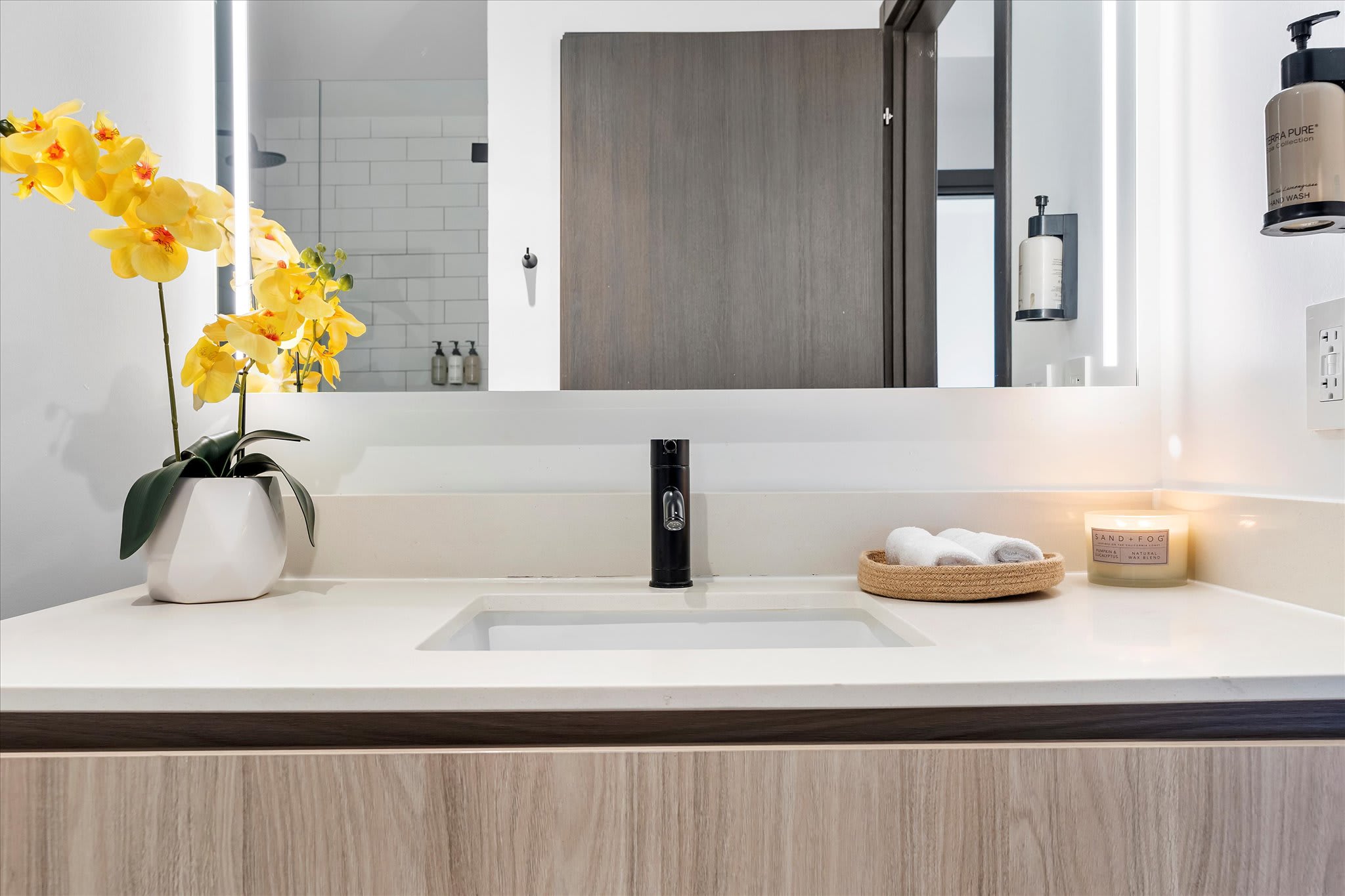 A close-up of a stylish bathroom vanity with a white countertop, integrated sink, and black faucet. It's adorned with vibrant yellow orchids, a woven basket with towels, and a scented candle.
