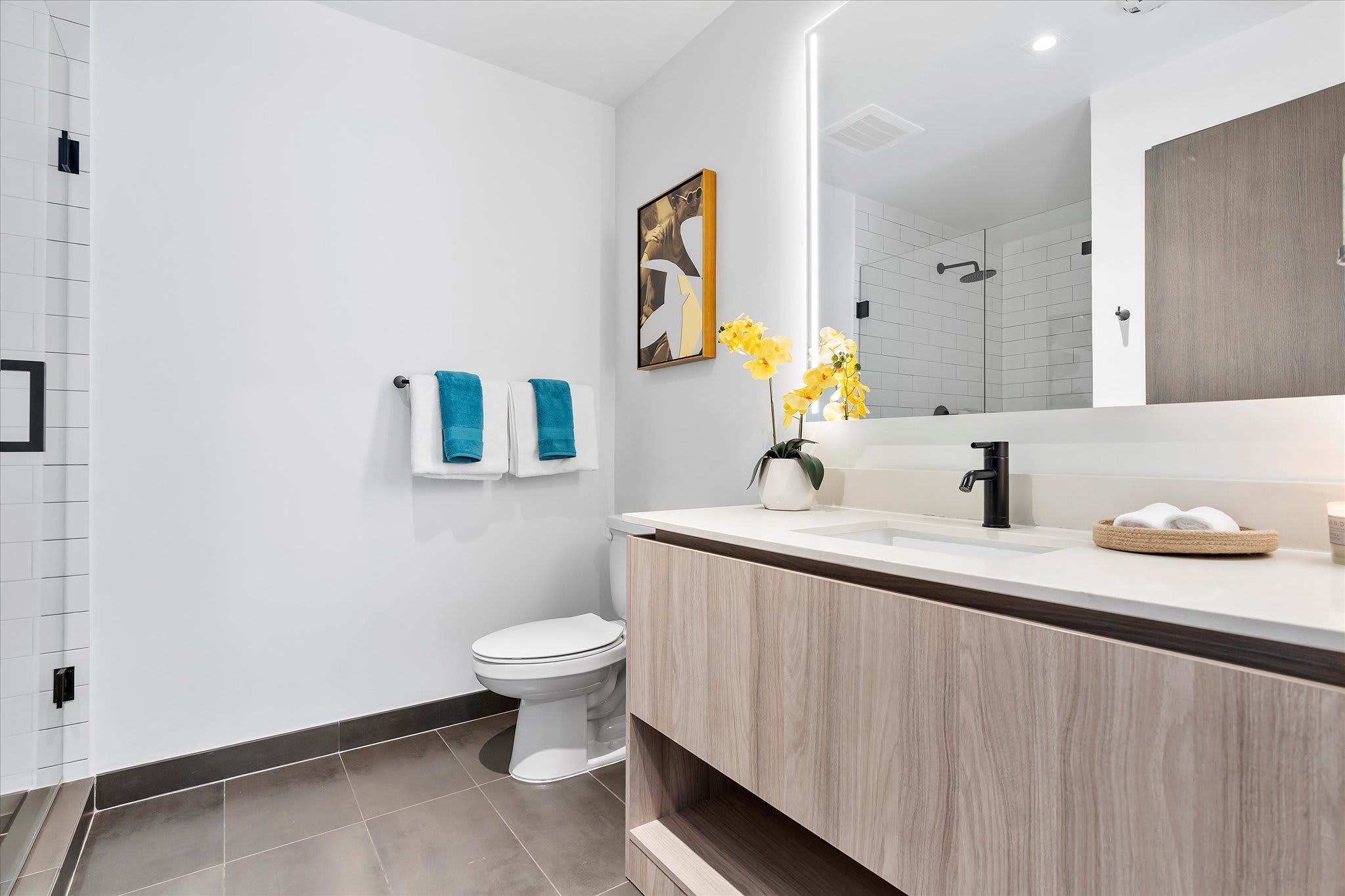 A bright and modern bathroom featuring a light wood vanity with a white countertop and black faucet, a toilet, and a walk-in shower with white subway tiles and a glass enclosure.