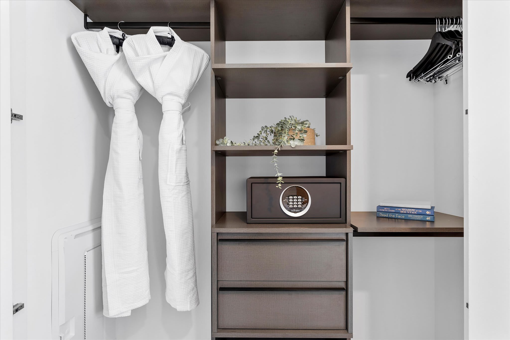 An organized closet area within the bedroom, featuring dark wood shelving, drawers, and hanging rods. Two white bathrobes, a secure safe, and decorative items are neatly arranged.