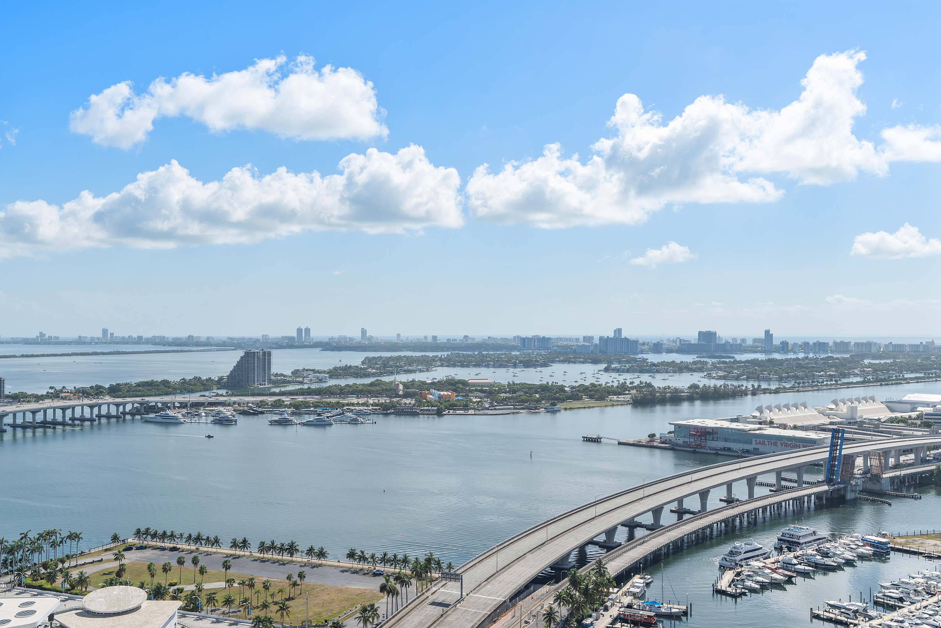 A breathtaking aerial view captures a sprawling cityscape, a wide bay dotted with numerous boats, and multiple bridges connecting landmasses under a bright blue sky with scattered clouds.