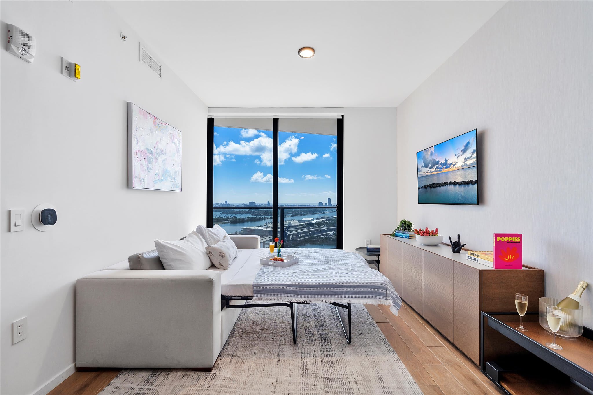 The living room transformed into a sleeping area with a sofa bed pulled out, featuring a wall-mounted TV, a long console, and a large window offering city and water views.