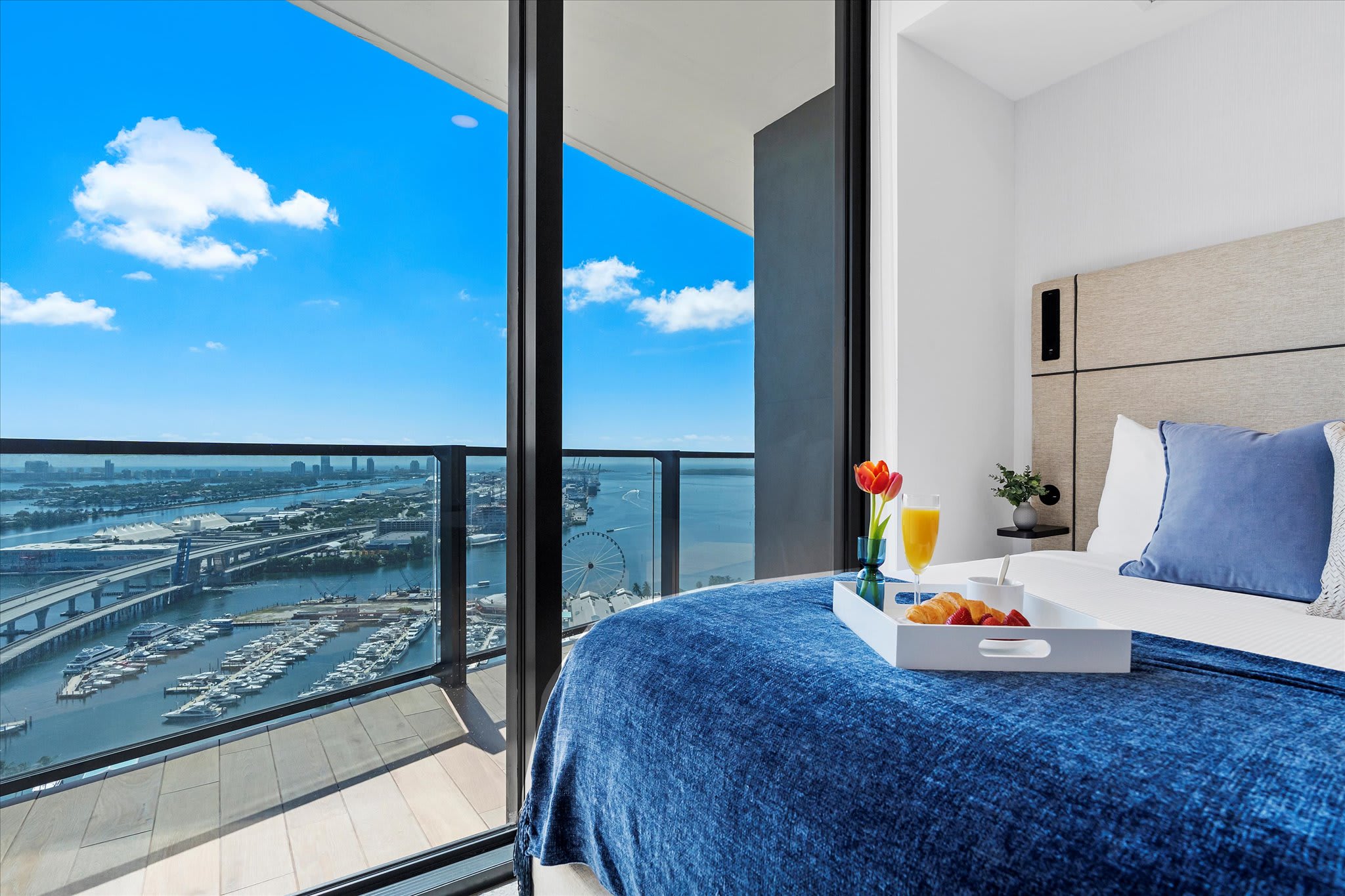 A cozy bedroom with a blue-covered bed and a breakfast tray, offering stunning panoramic views of the city skyline, marina, and ocean through a large floor-to-ceiling glass door leading to a balcony.