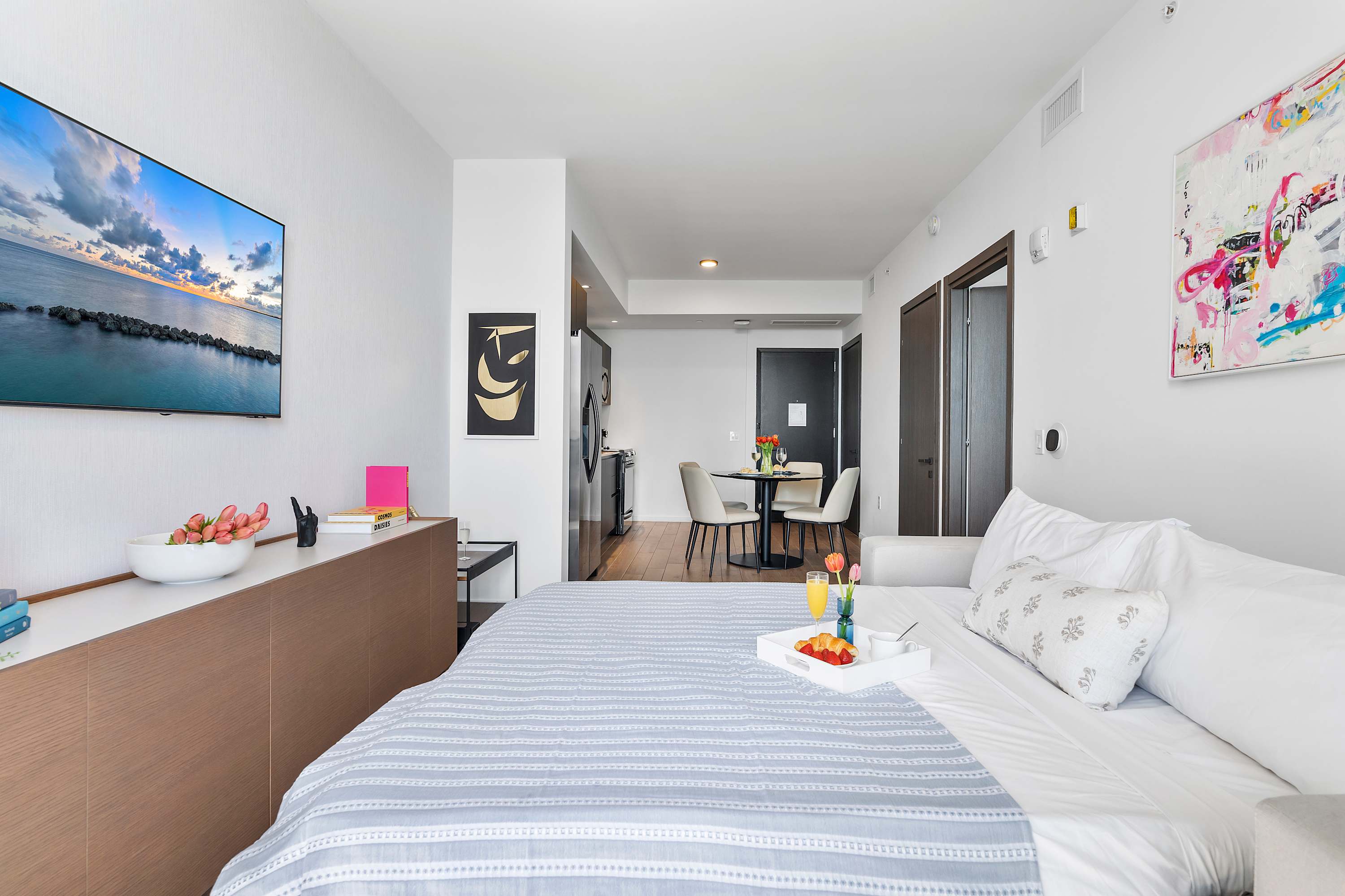An open-concept studio space featuring a bed with a breakfast tray, a small dining area with a round table and chairs, and a glimpse of the modern kitchen in the background, all under a bright ceiling light.