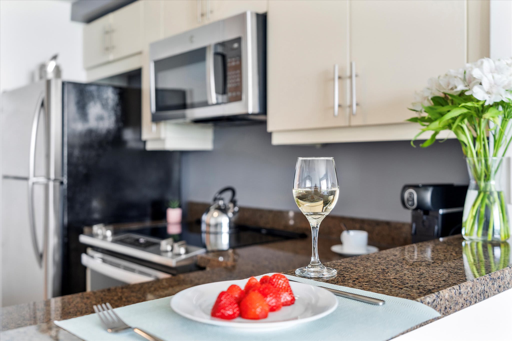 Modern kitchen featuring light beige upper cabinets, stainless steel appliances including a microwave and refrigerator, and dark granite countertops. A glass of white wine and a plate of fresh strawberries are neatly arranged on the counter.