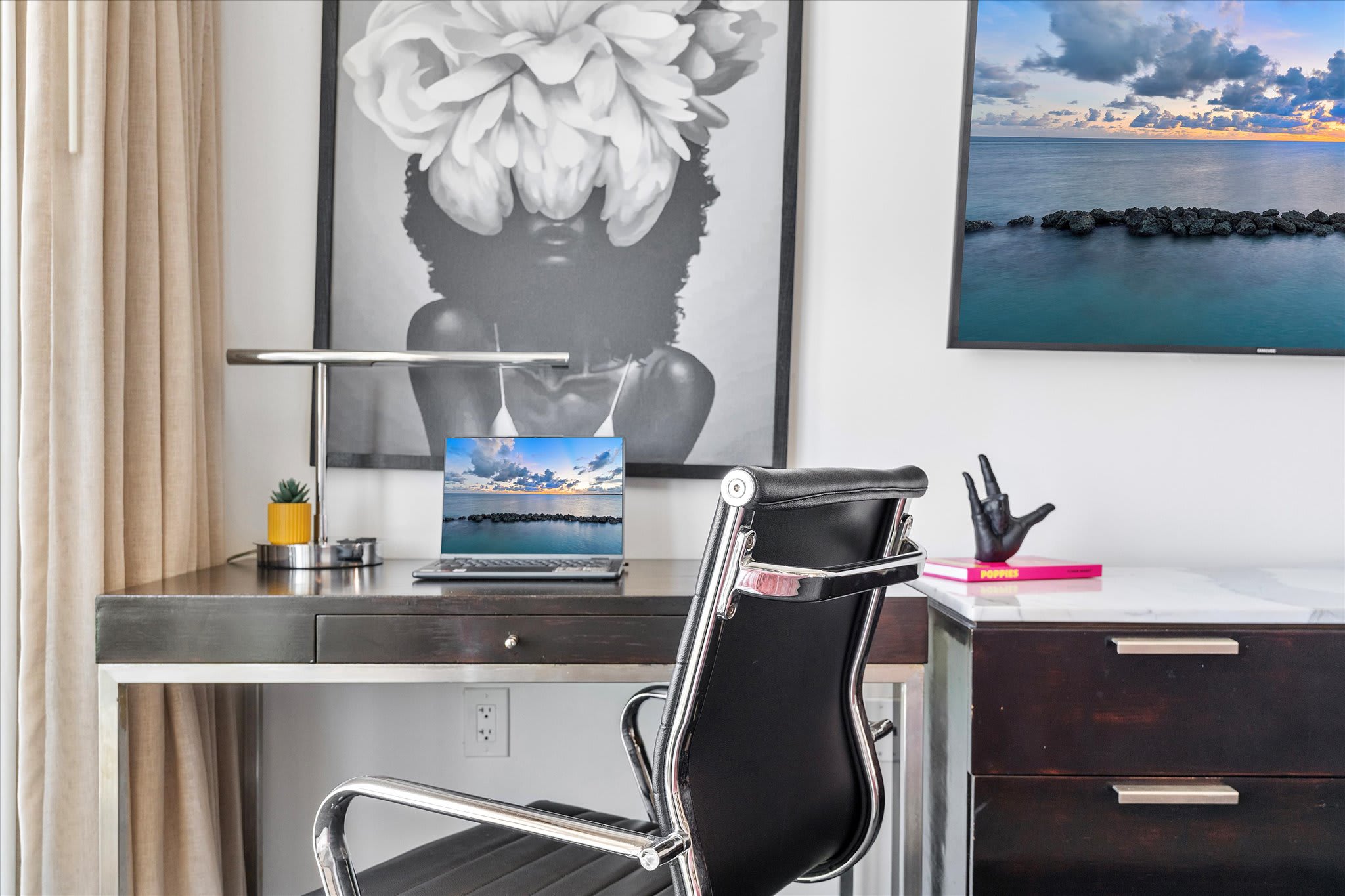 Detailed view of a modern workspace in the bedroom, featuring a sleek dark wood desk with a silver frame, a black office chair, and a laptop displaying a scenic view. A large black and white floral artwork hangs above the desk.
